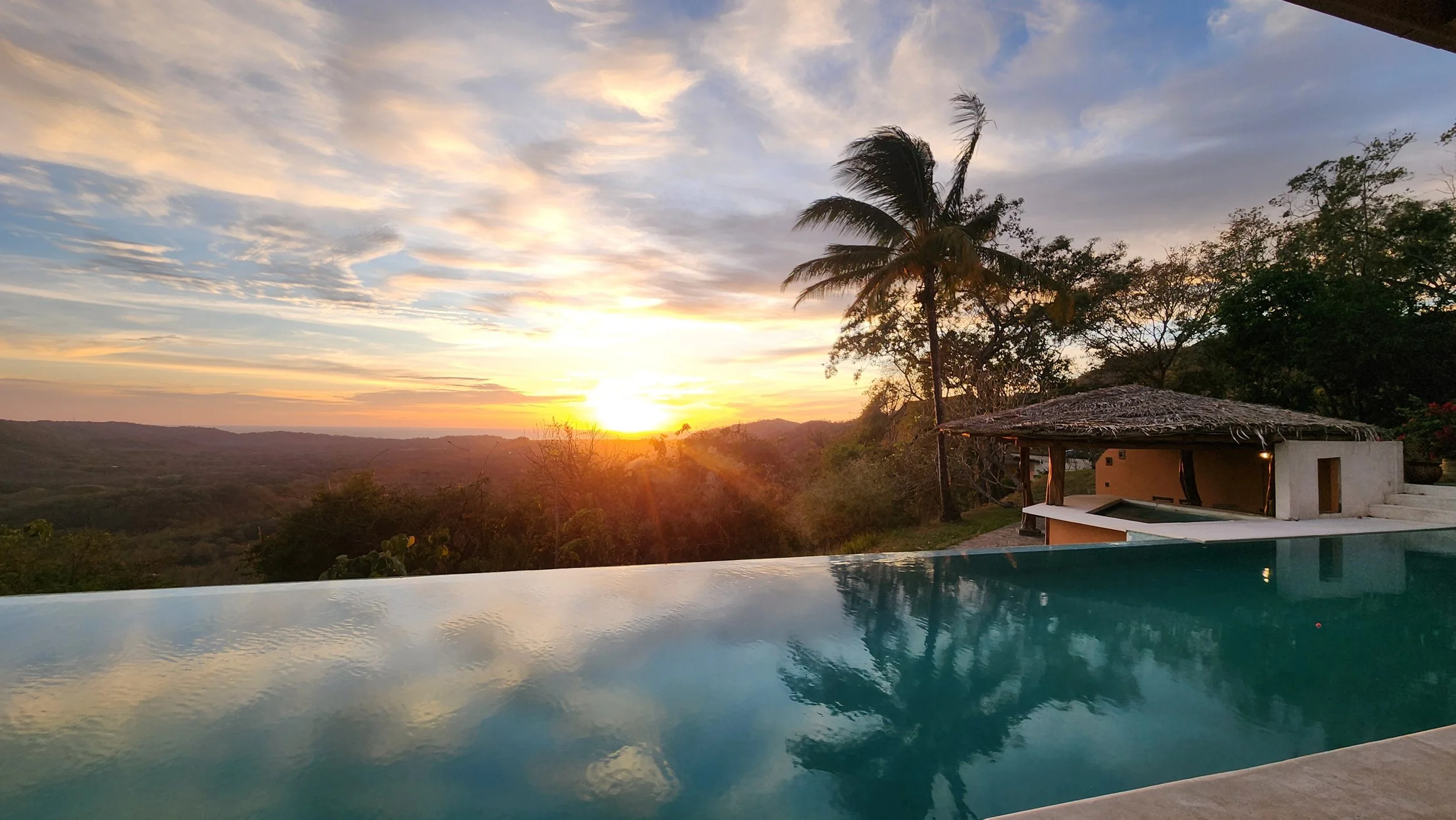 Sunset over a hilly landscape with a pool in the foreground, palm trees, and a thatched-roof structure at the playful soul's women's ayahuasca retreat.