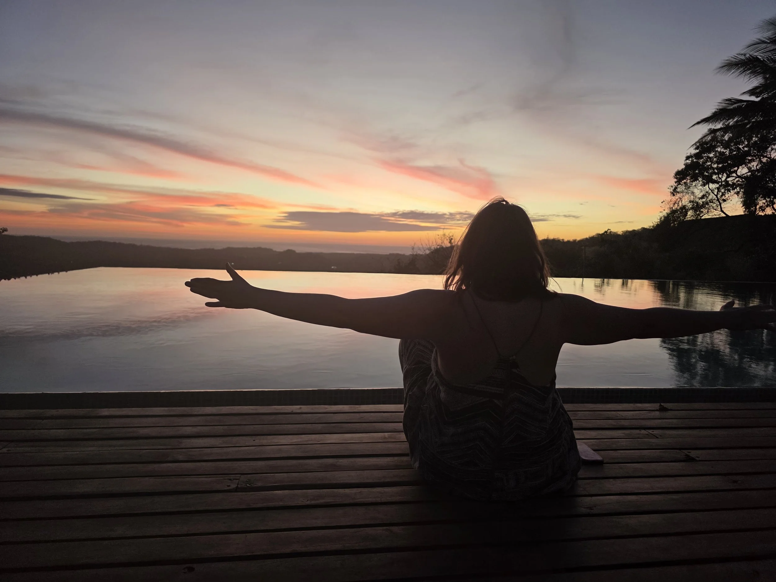 A woman at the women's ayahuasca reatreat with arms outstretched at sunset by the pool, with trees and clouds in the sky.