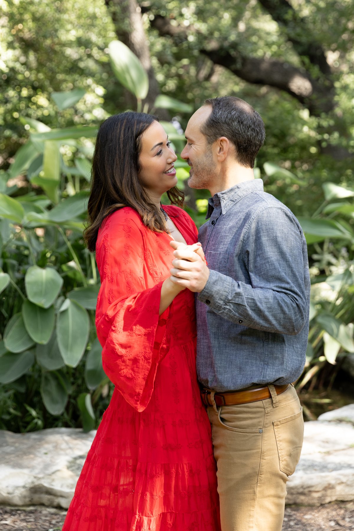 A couple dancing outdoors in a garden. The woman is wearing a red dress, and the man is wearing a blue shirt with khaki pants. They are holding hands and smiling at each other, surrounded by green foliage.