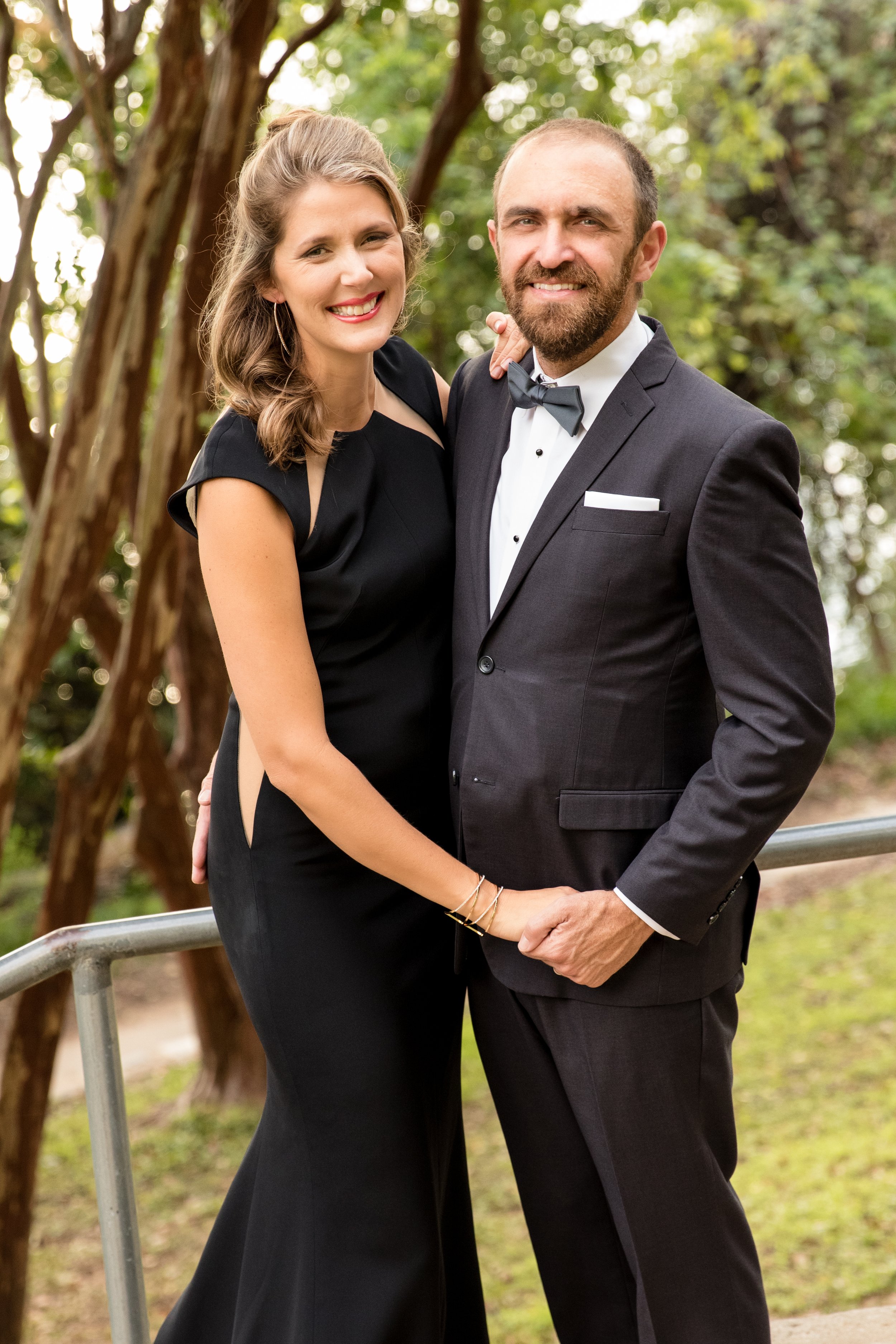 A smiling couple dressed in formal attire, with the woman in a black dress and the man in a suit with a bow tie, standing outdoors.