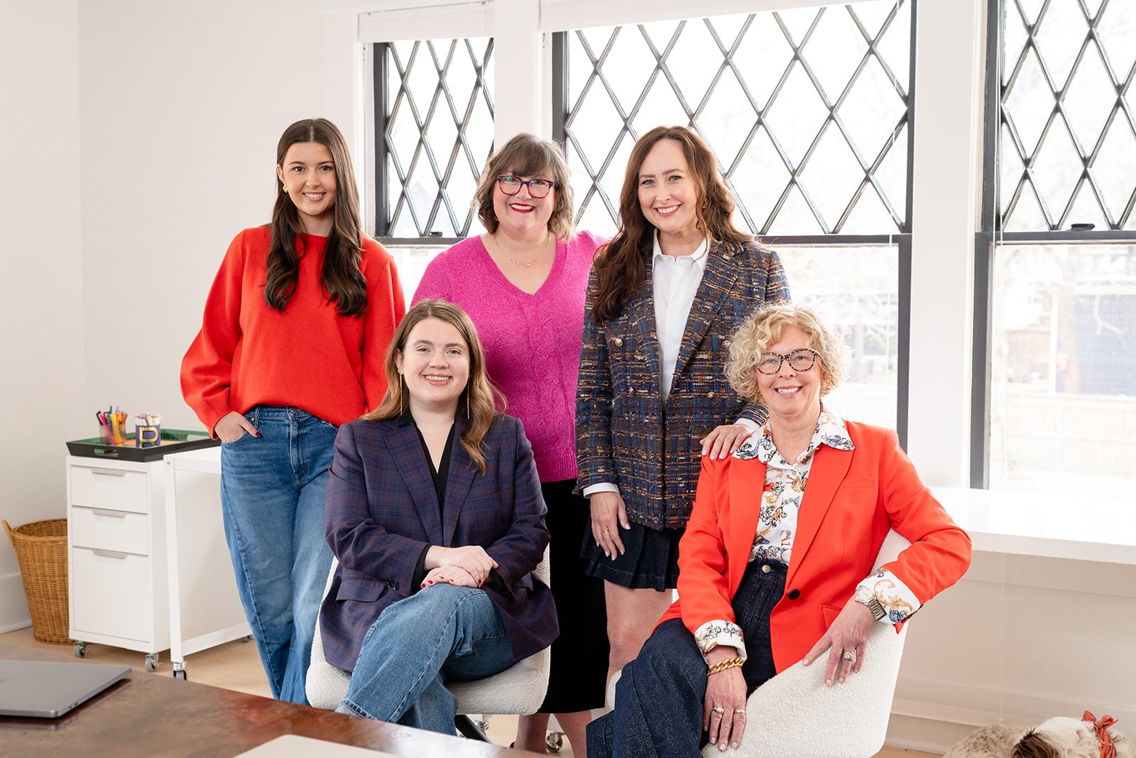 Group of five women of different ages and diverse styles posing together in an office or studio space with large windows and natural light.