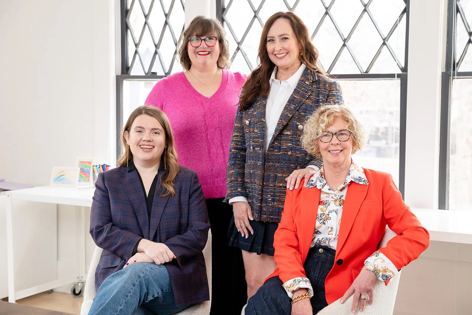 Five women posing in an office with large windows. One woman is sitting in a chair, and four women are standing behind her. They are all smiling.