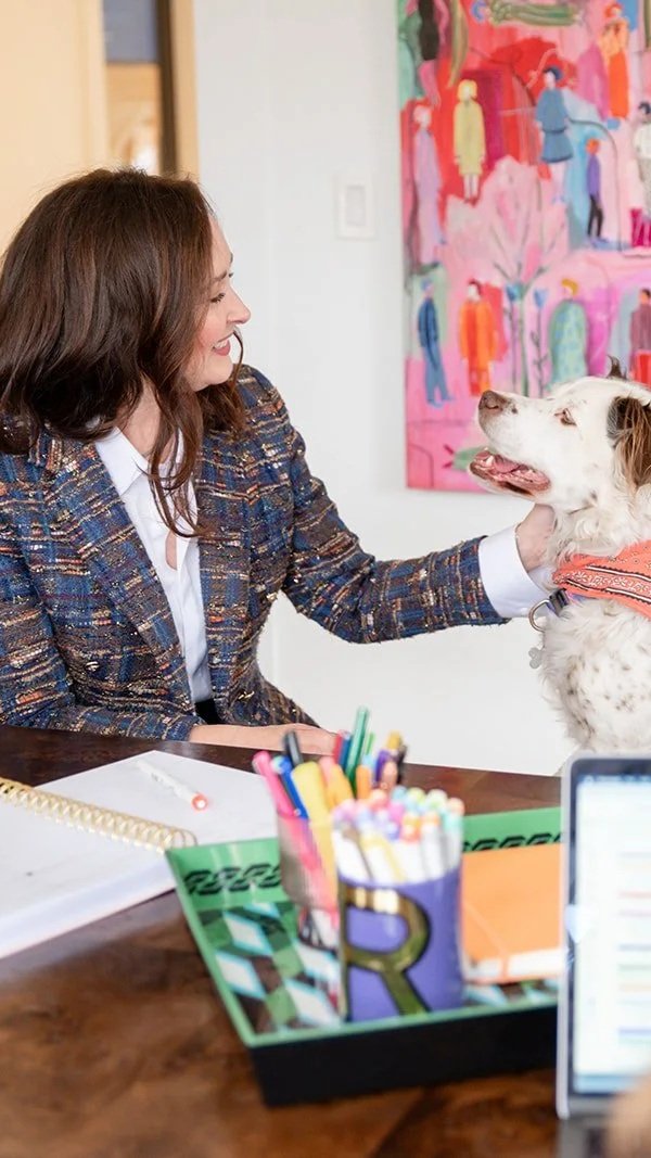 Woman smiling at her dog while petting it at a table in an office or meeting room, with colorful artwork on the wall in the background.