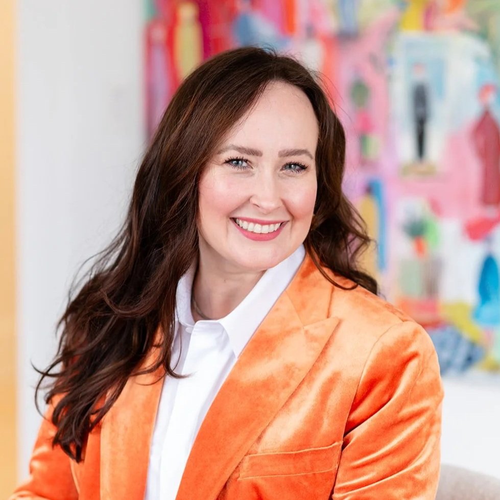 A woman with long brown hair wearing an orange blazer and white shirt, smiling in front of a colorful abstract artwork.