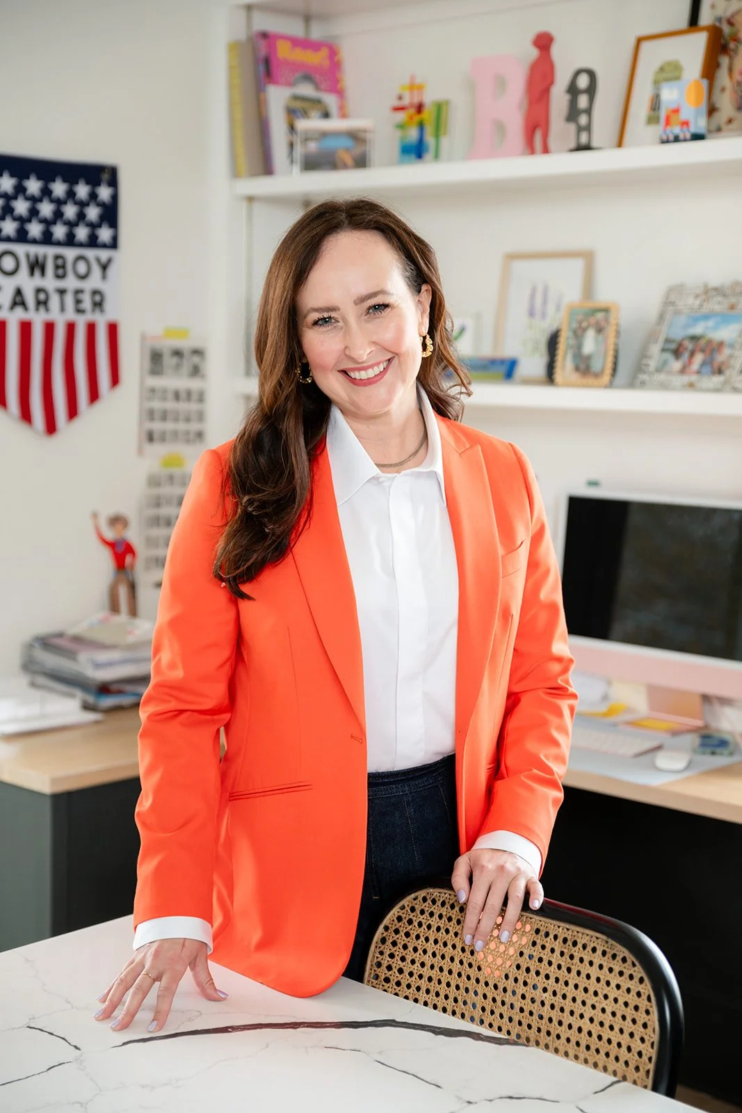 A woman with long brown hair, wearing a bright orange blazer and white shirt, smiling and standing behind a marble table in a colorful, decorated office.