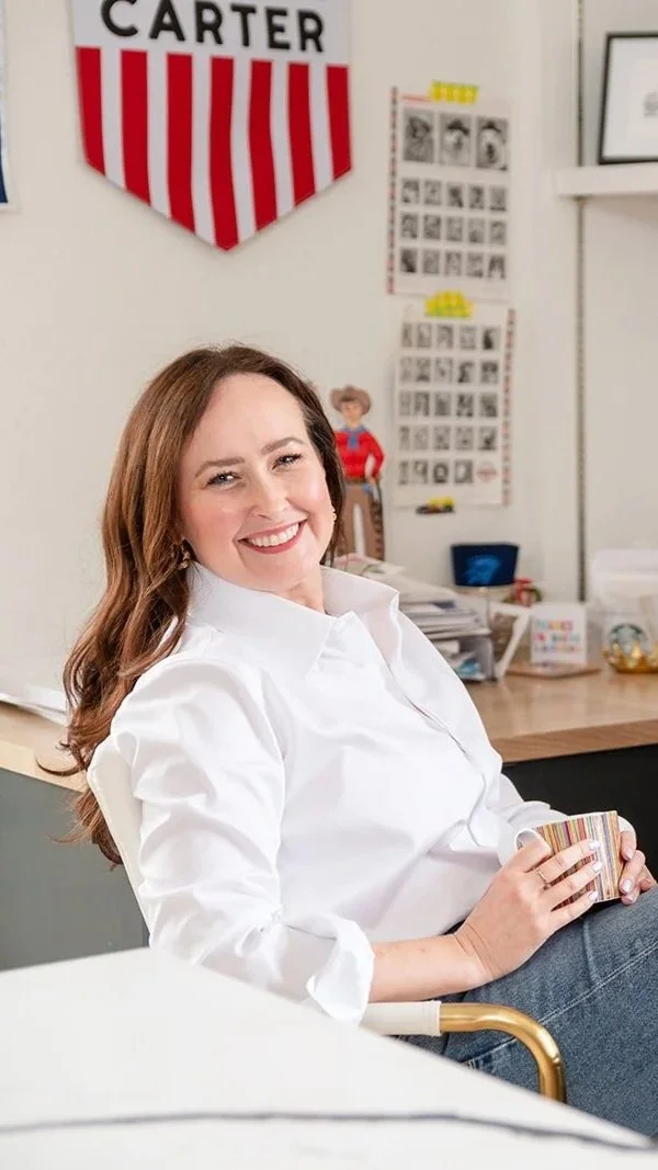 A woman with long auburn hair smiling and sitting in an office, holding a striped mug. Behind her are educational posters and a banner with red and white stripes reading "CARTER."