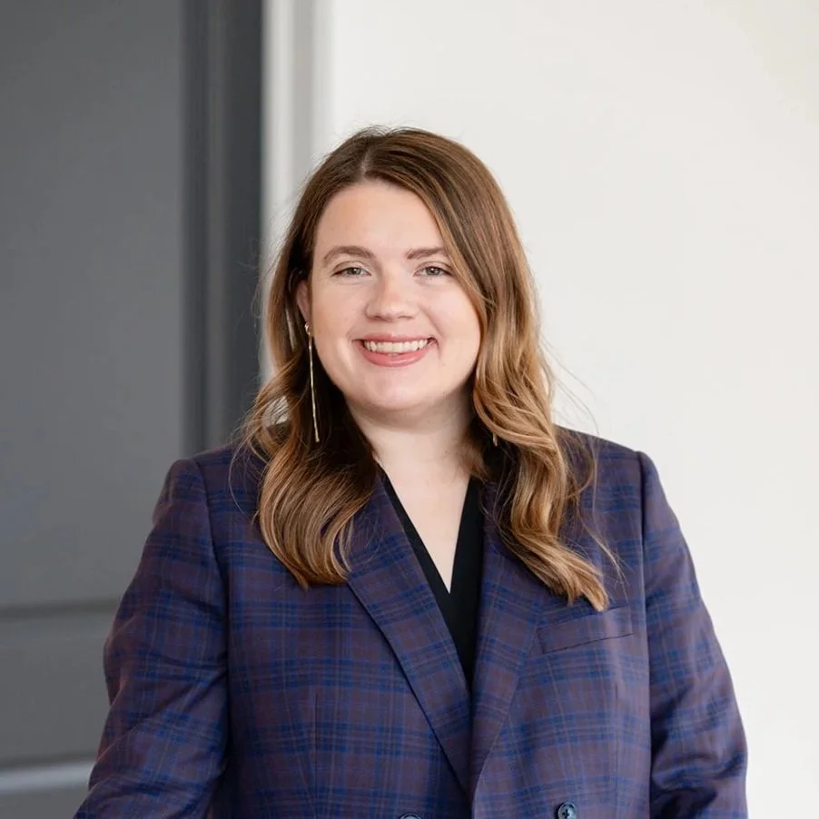 A woman with long, wavy auburn hair smiling, wearing a navy blue checked blazer and dangling earrings, standing indoors against a neutral background.