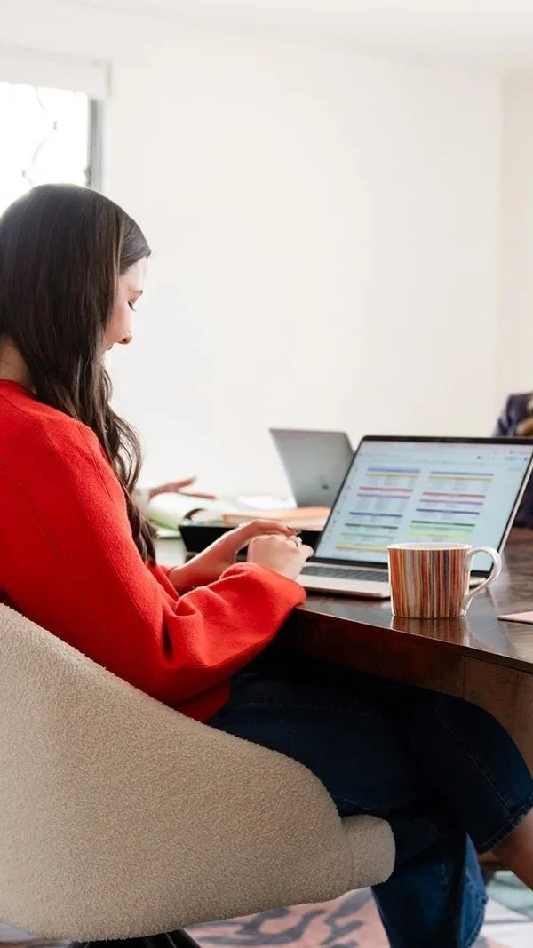 Woman in red sweater working on a laptop at a wooden table with a striped mug, in a bright room with a window.