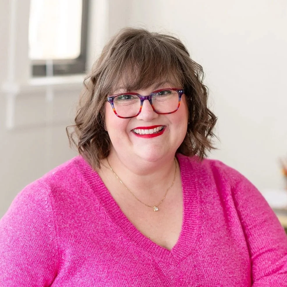 A woman with shoulder-length wavy brown hair, wearing colorful glasses, a pink sweater, and a gold necklace, smiling at the camera indoors near a window.
