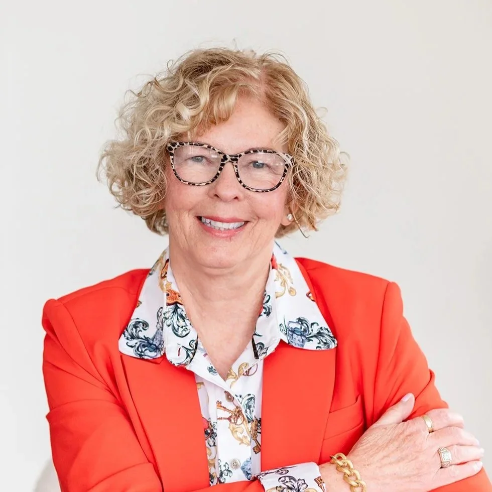 A smiling woman with curly blonde hair, wearing glasses with a leopard print frame, a white blouse with a colorful pattern, and a red blazer. She has her arms crossed and is posing against a plain white background.