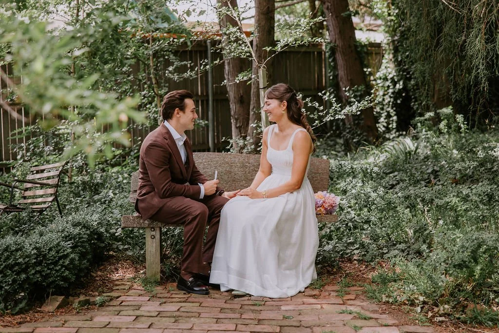 A man in a brown suit and a woman in a white dress sitting on a bench in a garden, holding hands and smiling at each other.