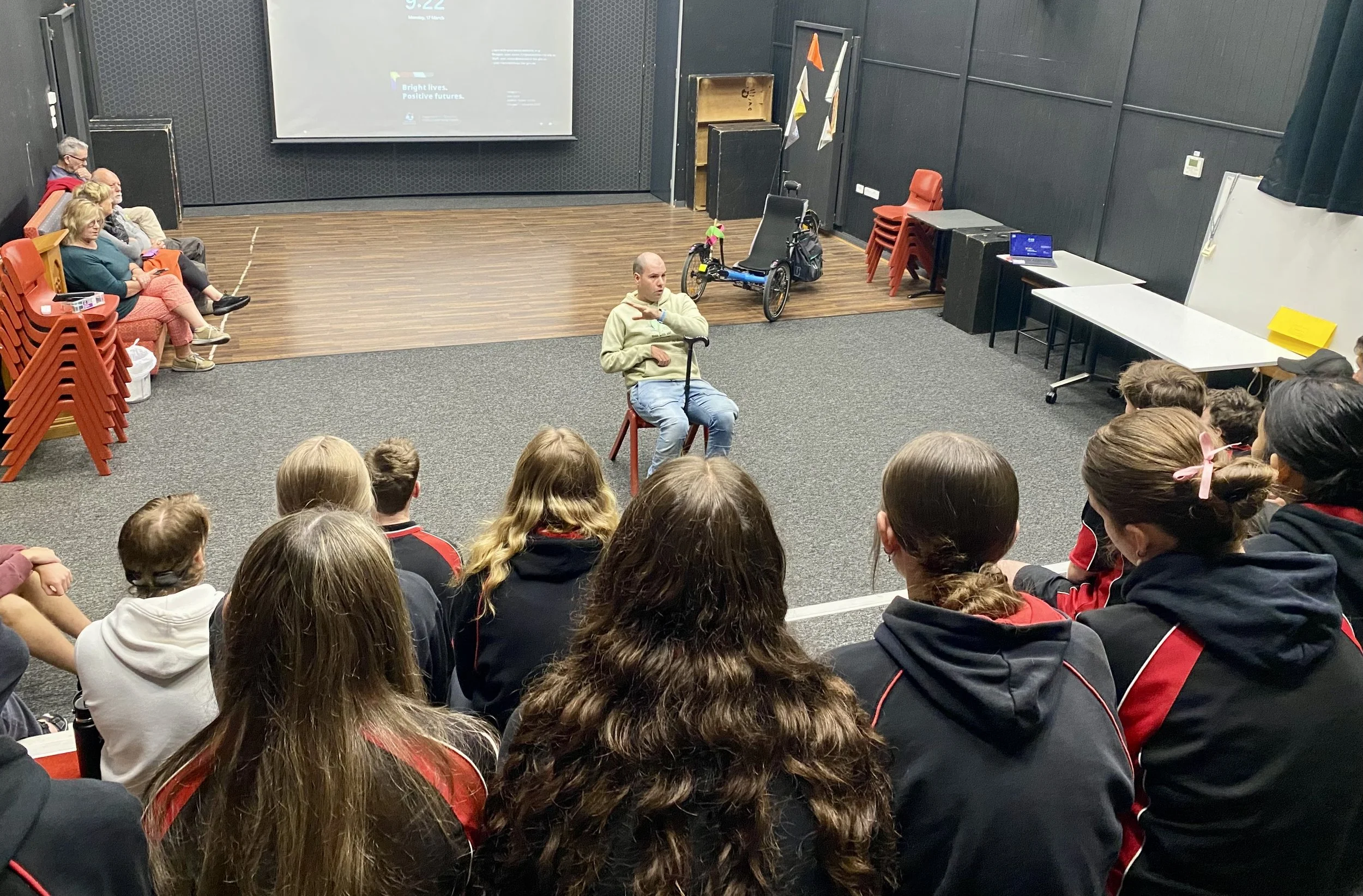 A group of teenagers and adults are seated on chairs in a room with a large projector screen and dark walls, watching a presentation or speech. One man is sitting on a chair in the middle, gesturing with his hand. A trike is behind him.