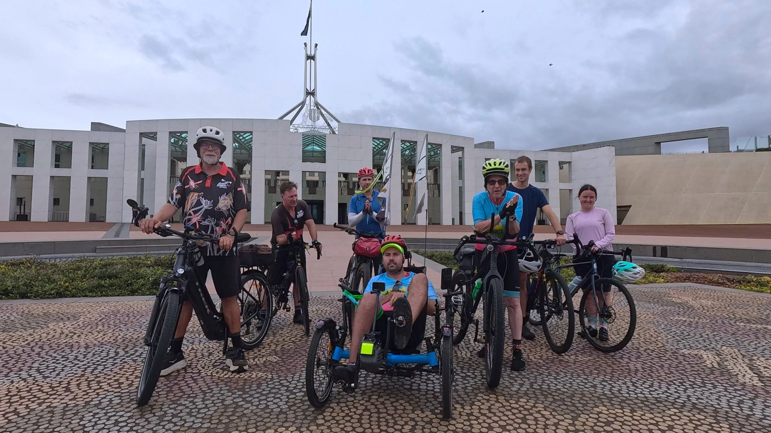 Group of seven people, including six with bicycles and one with a recumbent trike, posing in front of the government house building with flags, on a cloudy day.