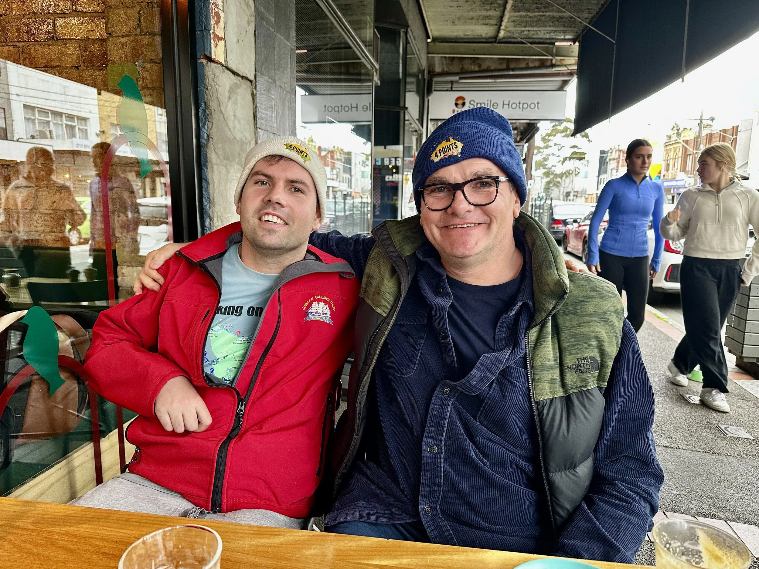 Two men sitting at an outdoor cafe table, wearing beanies, smiling at the camera, with several women and cars in the background on a city street.