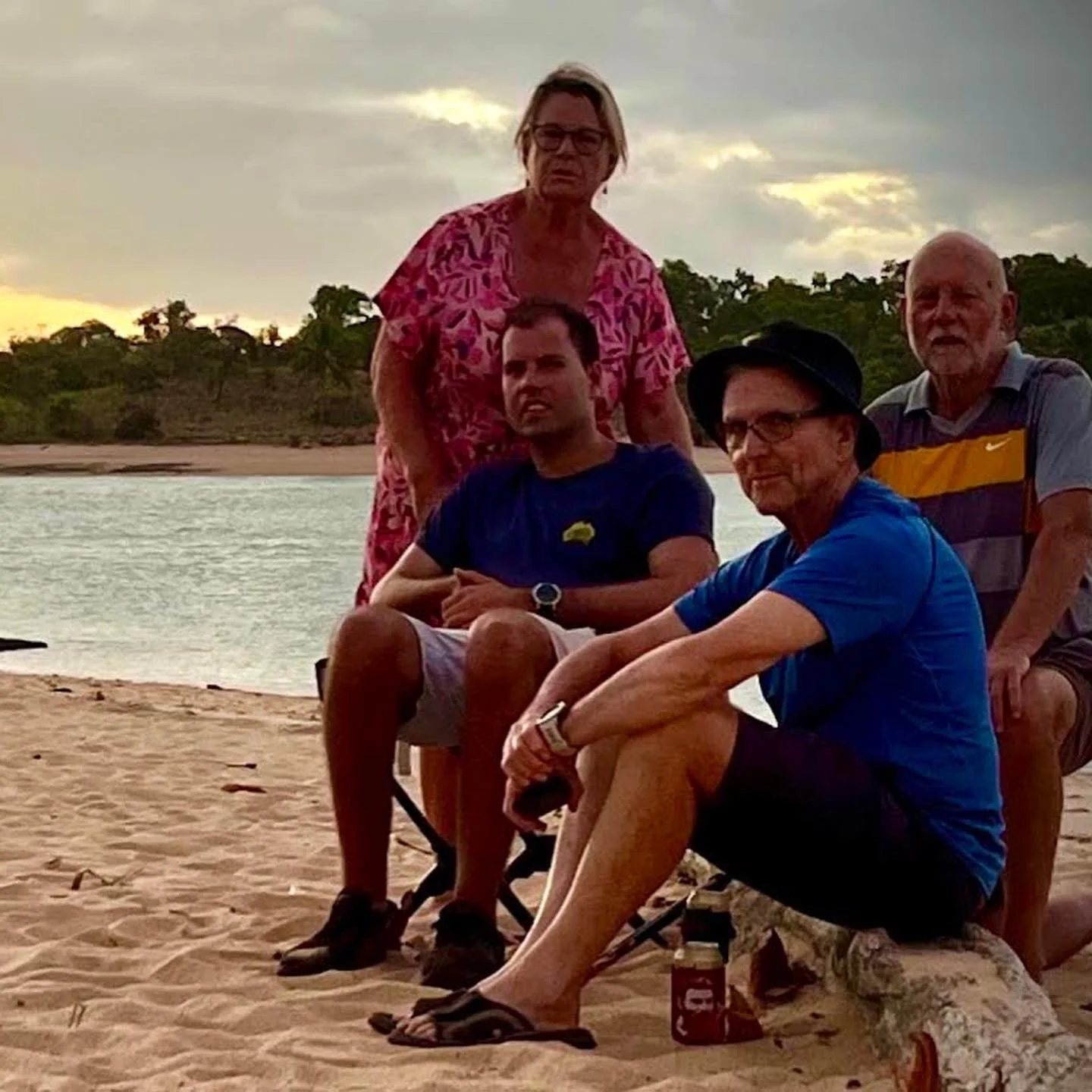 Four people on a beach near water, posing for a photo during sunset. Three men are seated on or near a log, while a woman in a pink shirt stands behind. The scene is outdoors with trees and a cloudy sky in the background.