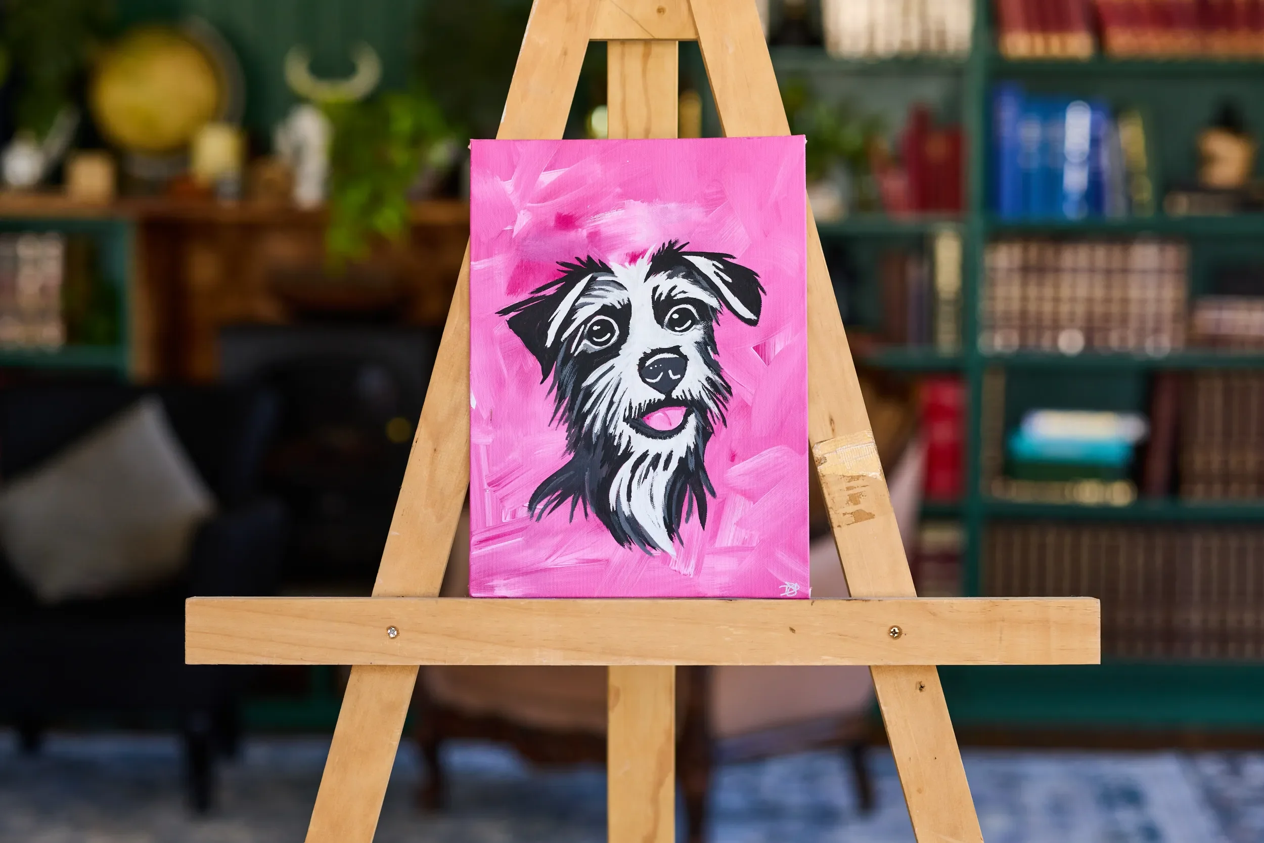 Painting of a scruffy black and white terrier on a hot pink canvas, centred on a wooden easel in front of green bookshelves.