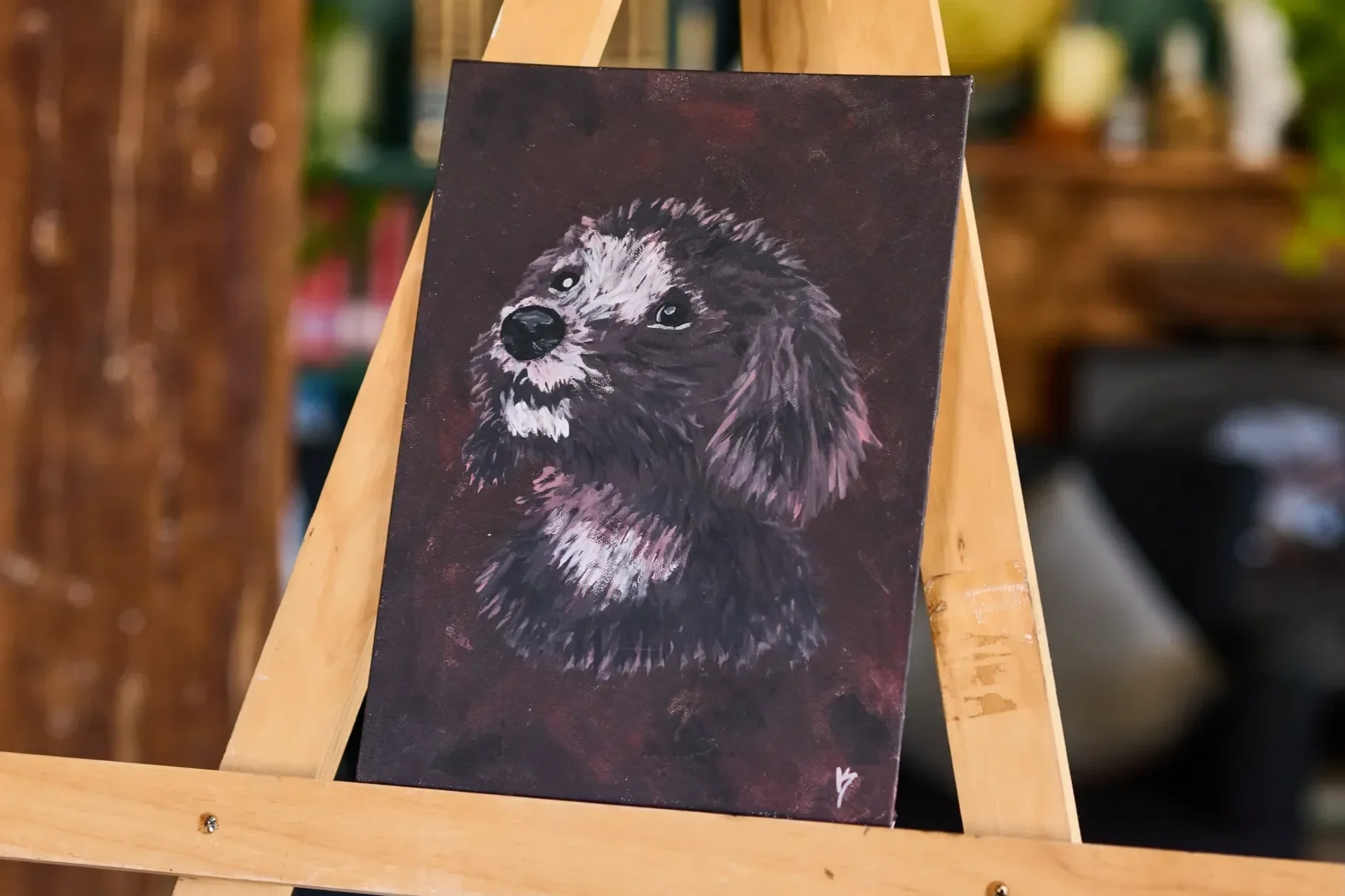 Painting of a dark brown shaggy dog looking up and to the left on a deep maroon canvas, resting on a wooden easel