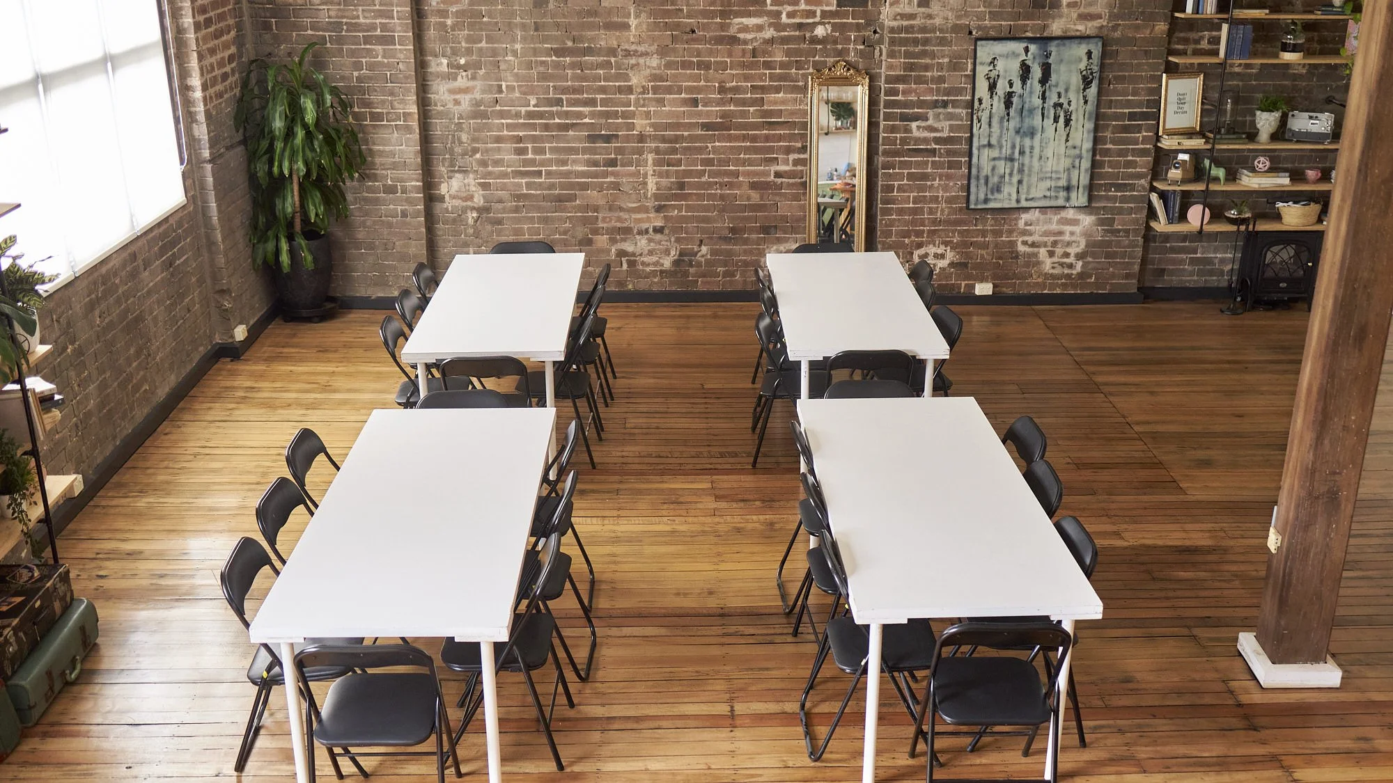 Industrial-style meeting room with four white tables, black folding chairs, exposed brick walls, wood floors, plants, artwork, and shelving