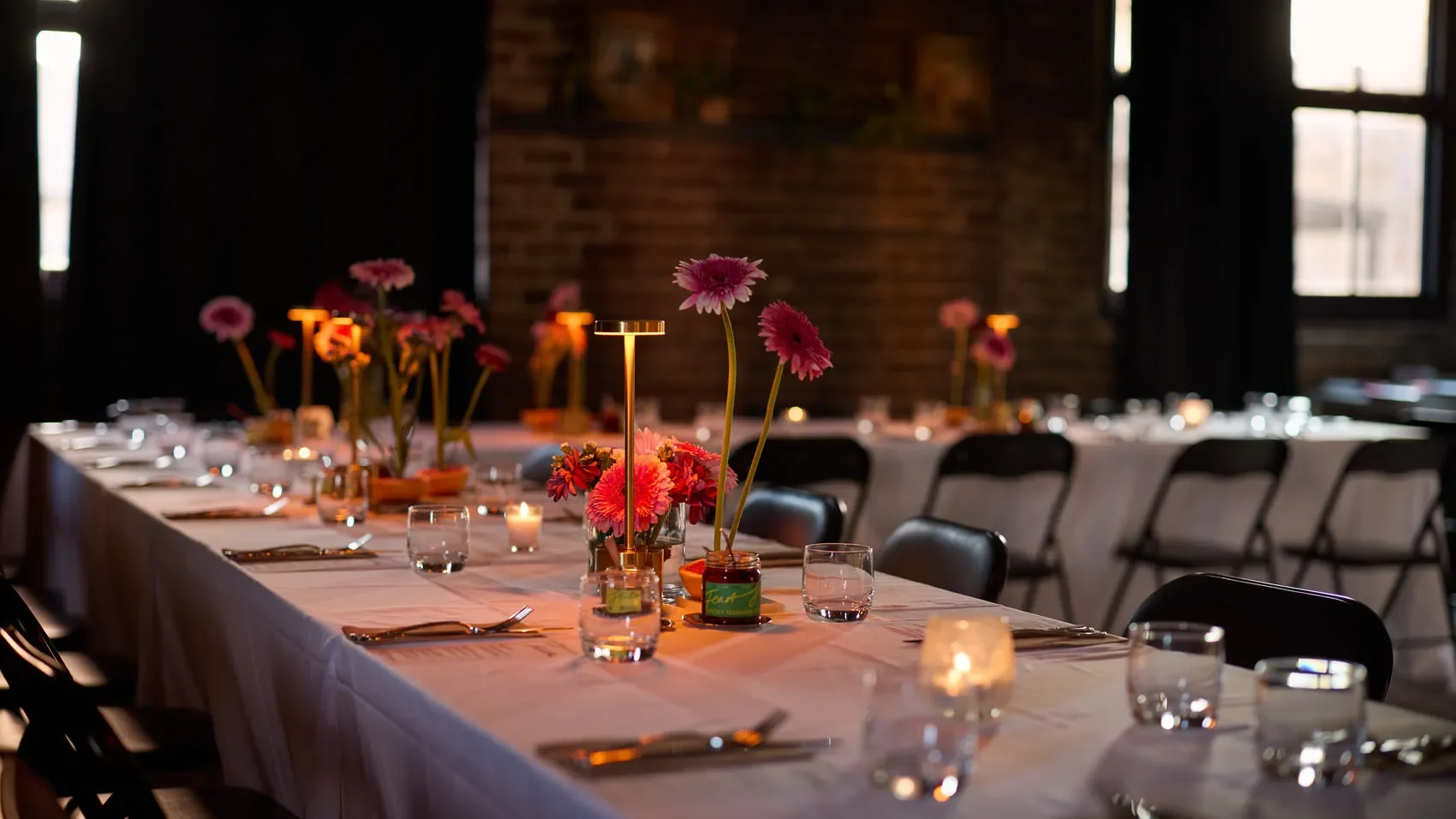 Elegant dinner table with white linens, floral centrepieces, candles, place settings, and black chairs in a brick-walled venue at the Dale Library in Chippendale. 