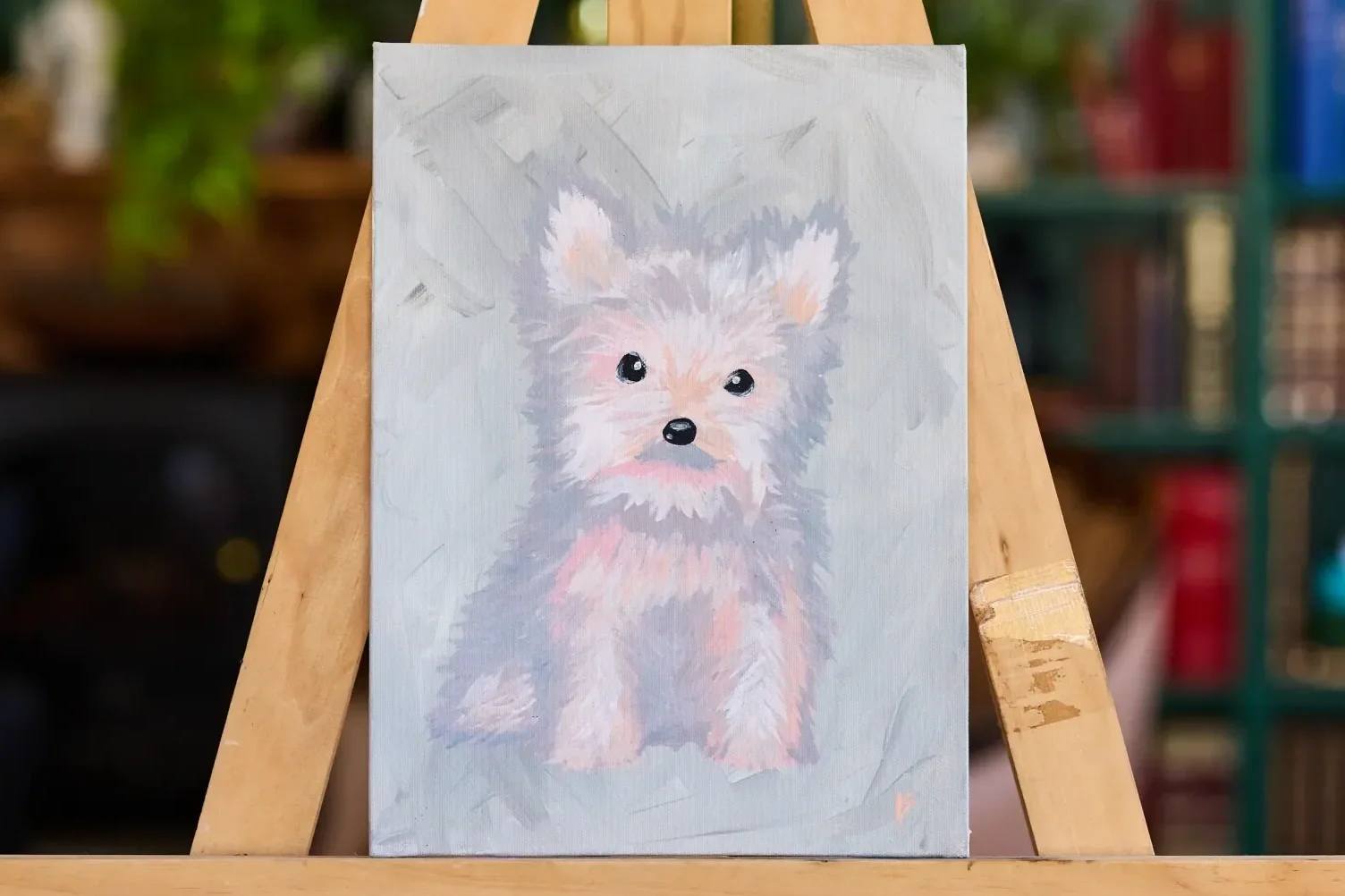 Painting of a grey and pink Yorkshire terrier puppy sitting on a pale grey canvas, on a wooden easel in front of bookshelves.