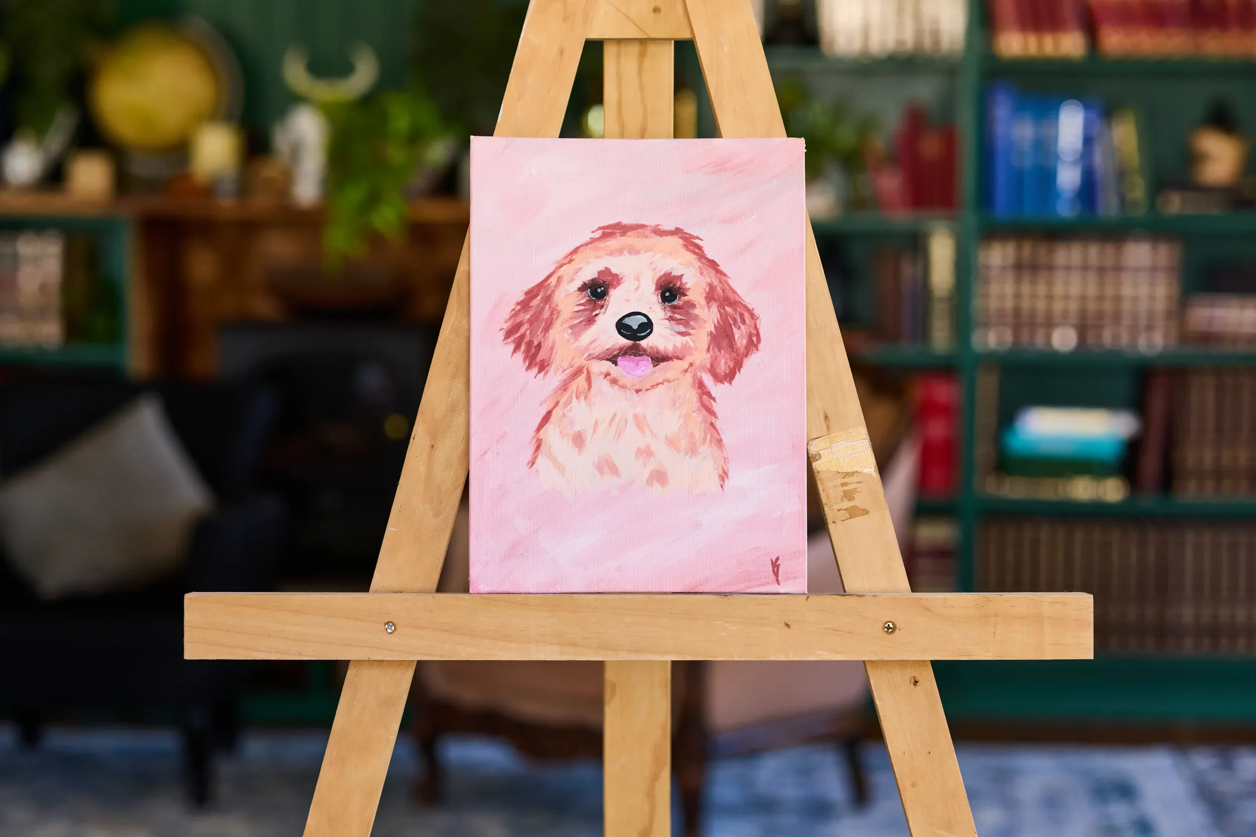 ainting of an apricot curly-coated puppy with its tongue out on a pale pink canvas, on a wooden easel in a library room.