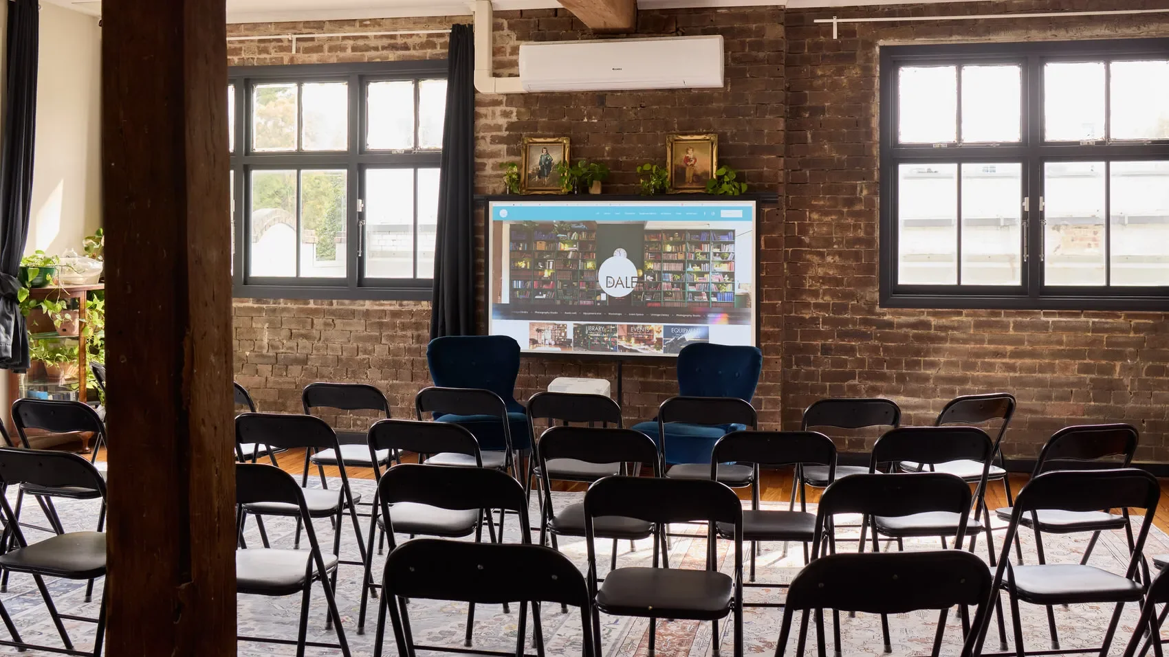 Industrial brick-walled event room with rows of black folding chairs, blue lounge chairs, and a large screen displaying a website at the Dale Library in Chippendale. 