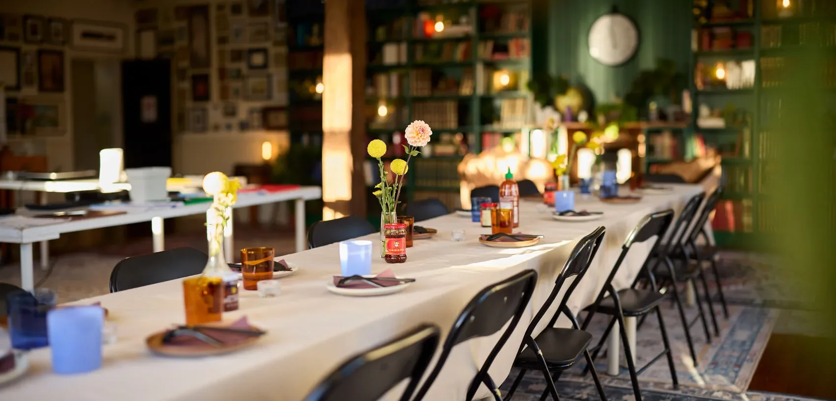 Long communal dining table set with plates, candles, flowers, and black chairs in a warmly lit room with bookshelves at the Dale Library in Chippendale. 