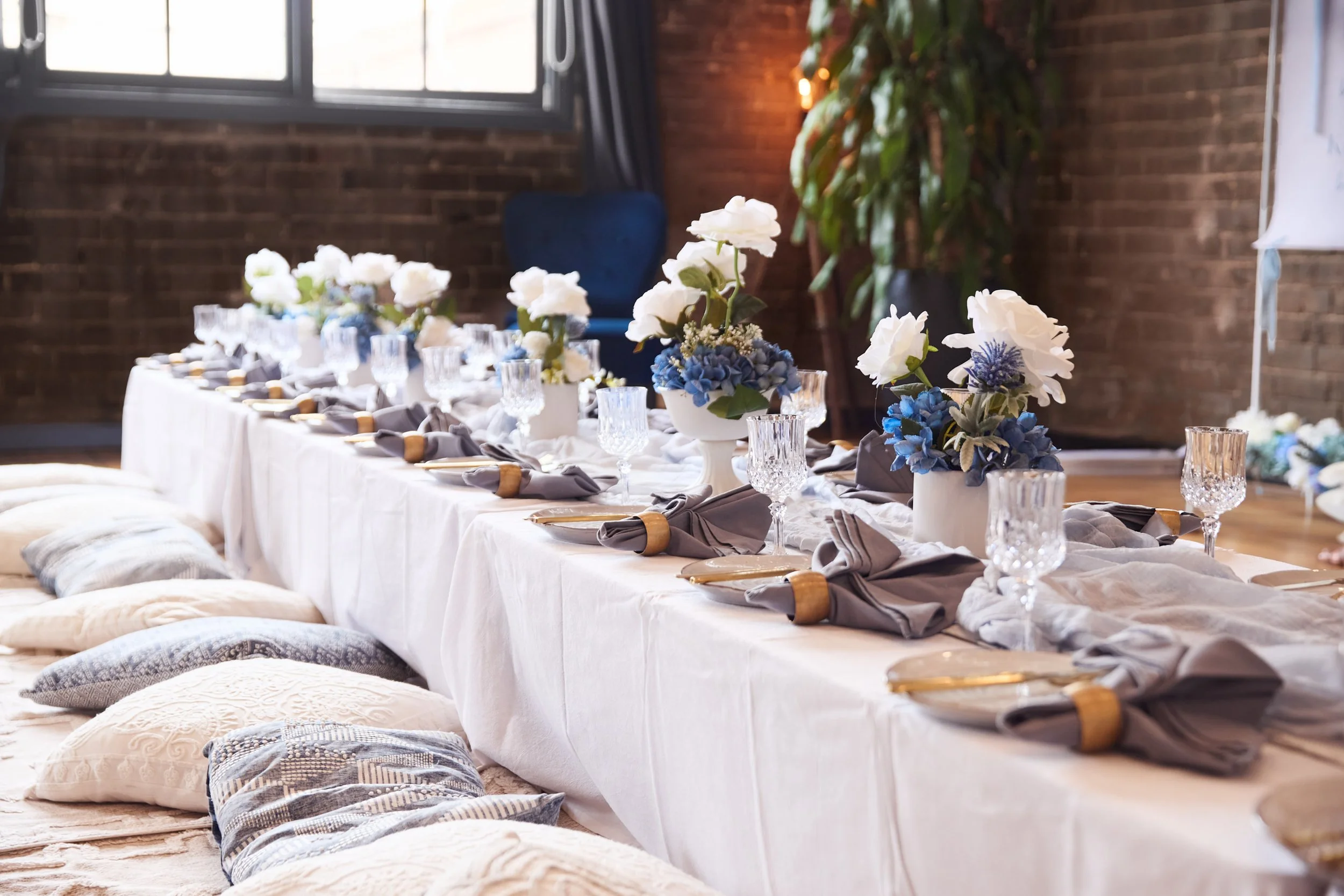 Elegant white-and-blue floral tablescape with gray napkins, gold flatware, crystal glassware, and floor cushions in a brick-walled venue