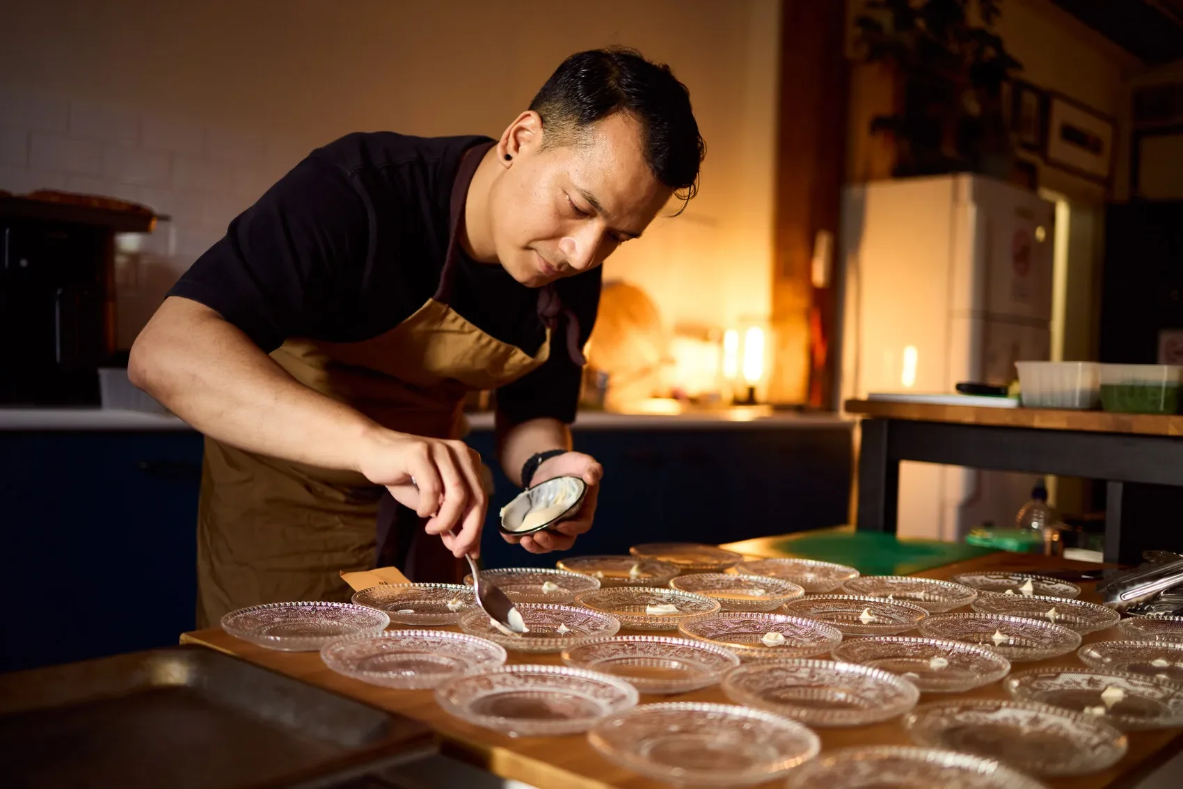 Chef in an apron spooning cream onto multiple glass dessert plates in a kitchen