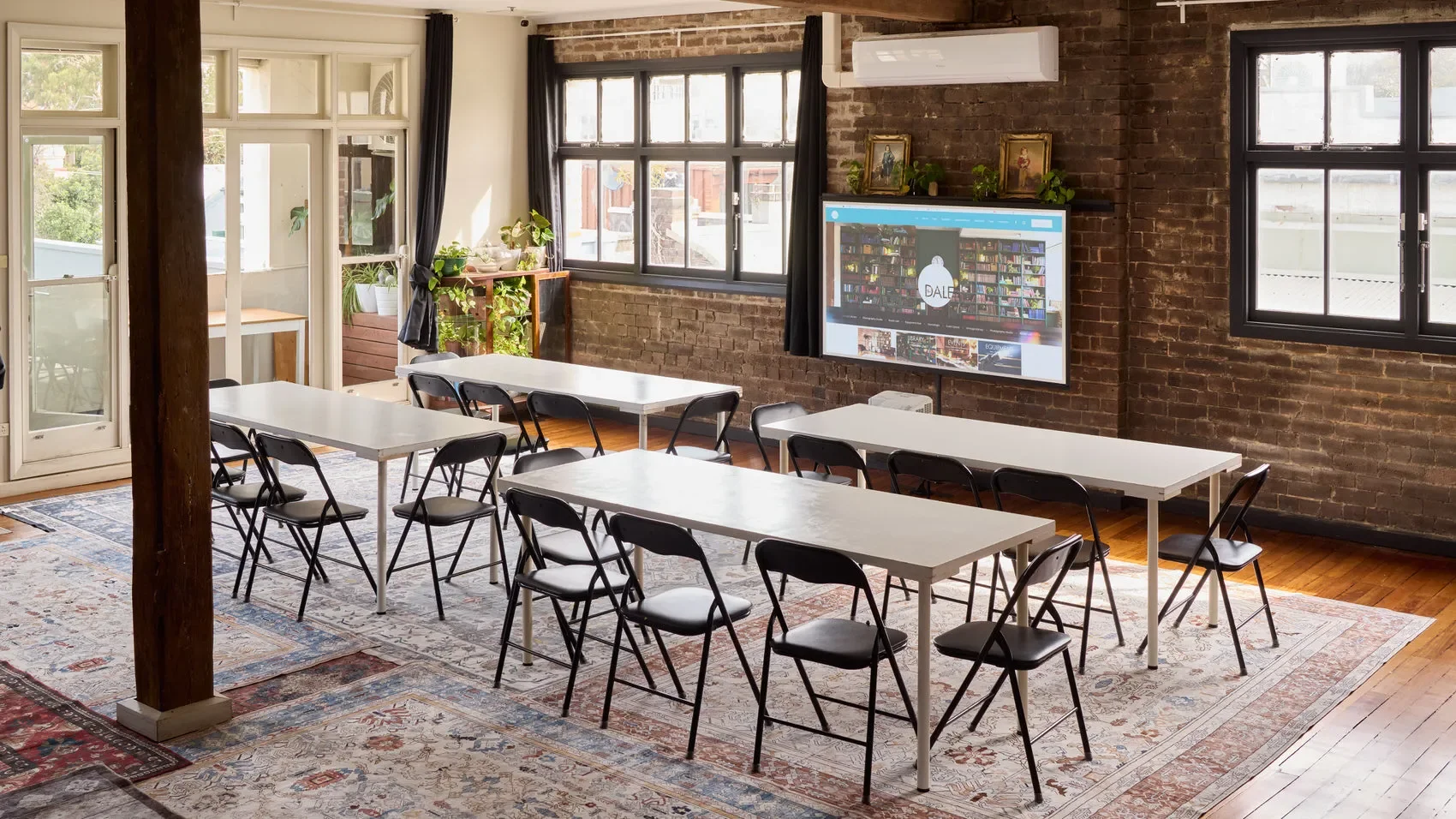 Bright brick-walled meeting room with white tables, black folding chairs, indoor plants, large windows, and a wall-mounted display at the Dale Library in Chippendale. 