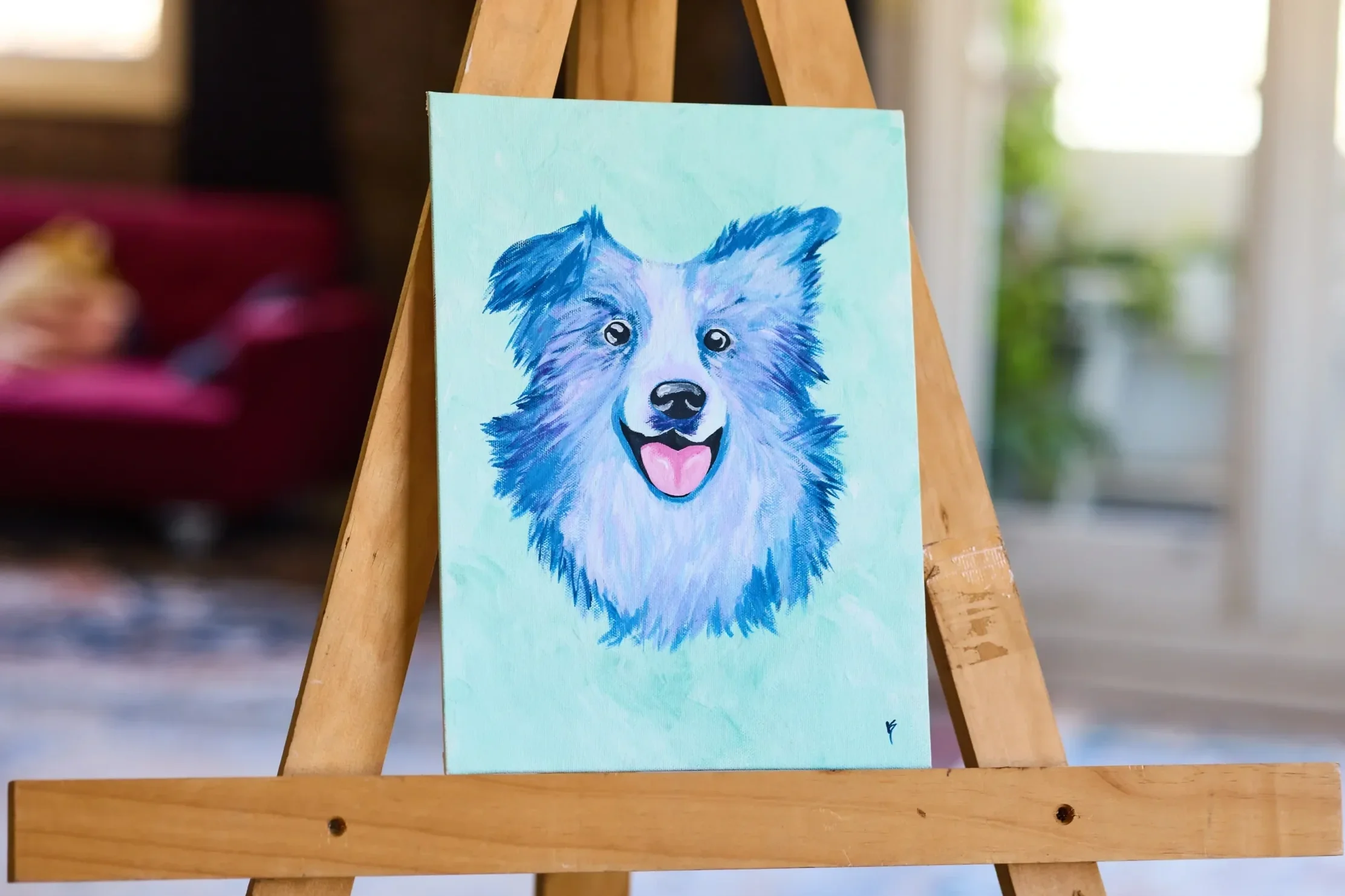 Close view of a blue and lilac collie painting on a mint canvas, on an easel in a bright room with a red sofa behind.