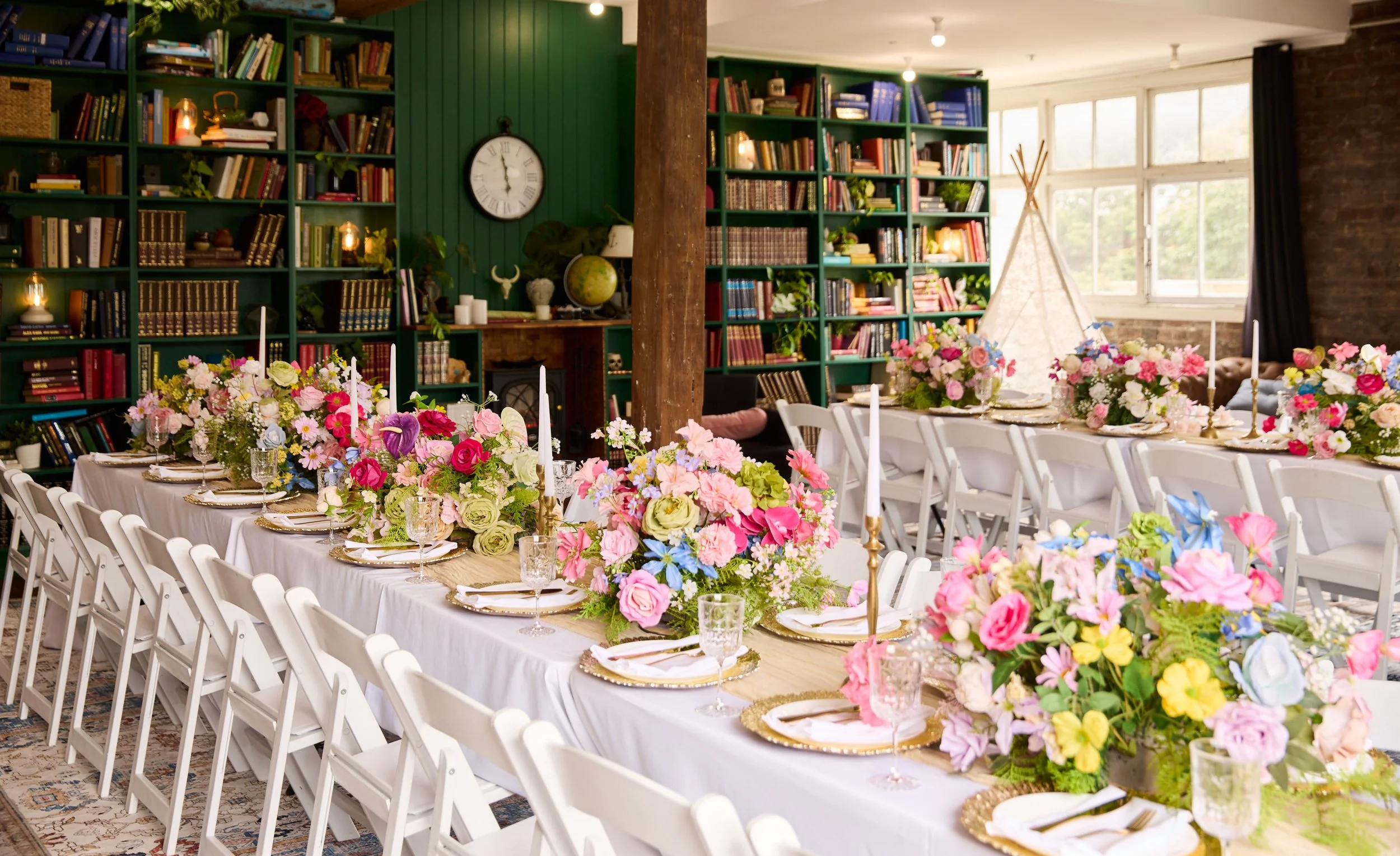 Elegant event dining room with long white-clothed tables, colorful floral centerpieces, gold-rimmed place settings, candles, white folding chairs, and green bookshelves at the Dale Library in Chippendale. 