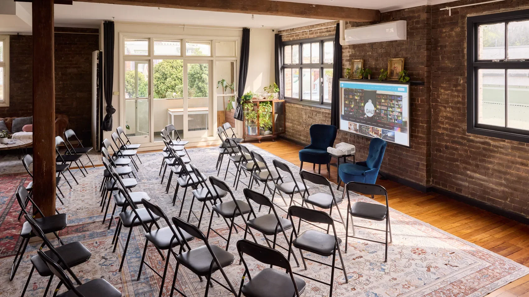 Loft-style event room with rows of folding chairs, projector, blue armchairs, brick walls, large windows, and indoor plants at the Dale Library in Chippendale. 