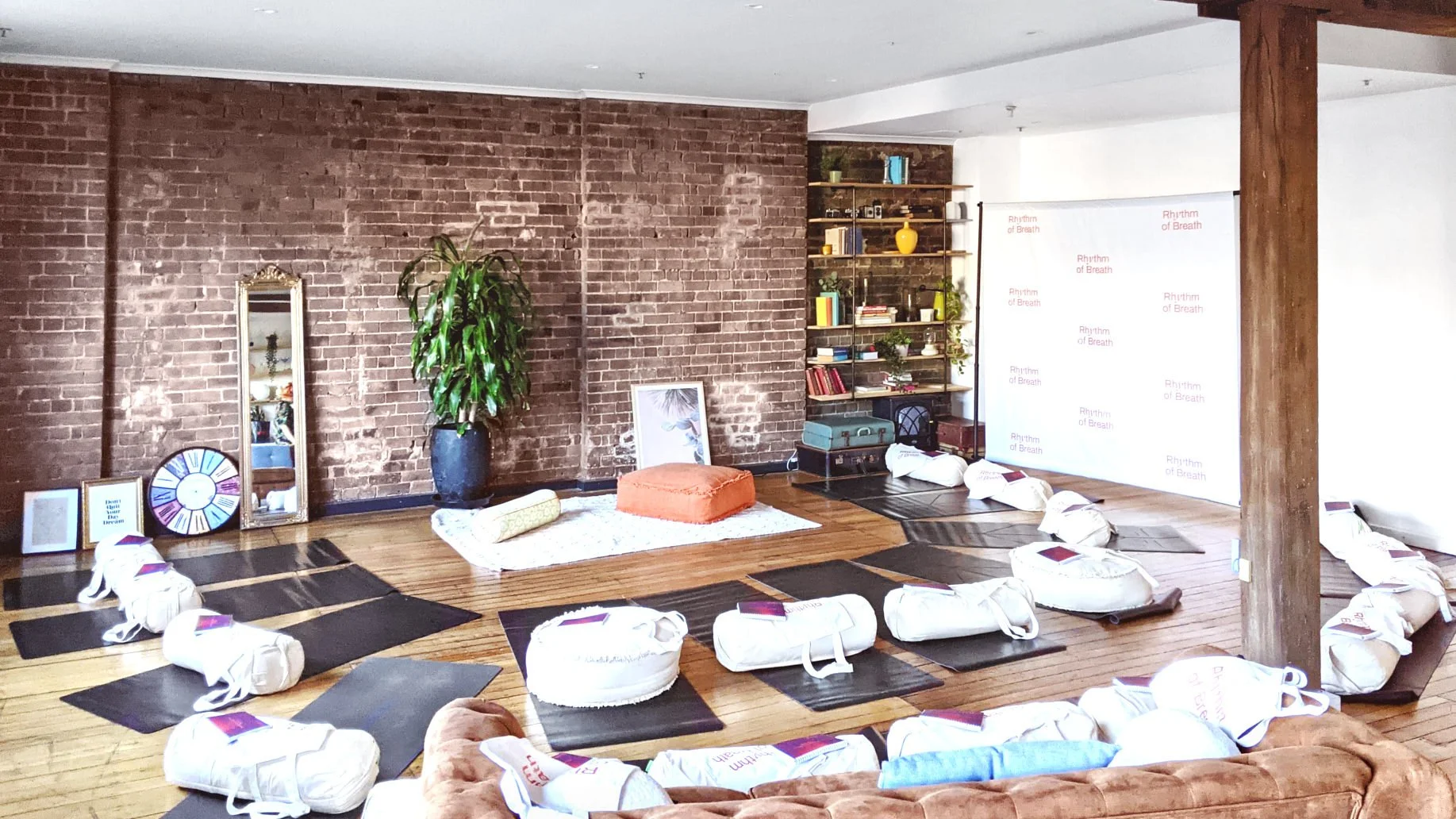 Yoga studio with mats, white meditation cushions, bolsters, brick walls, plants, and a screen displaying “Rhythm of Breath.”