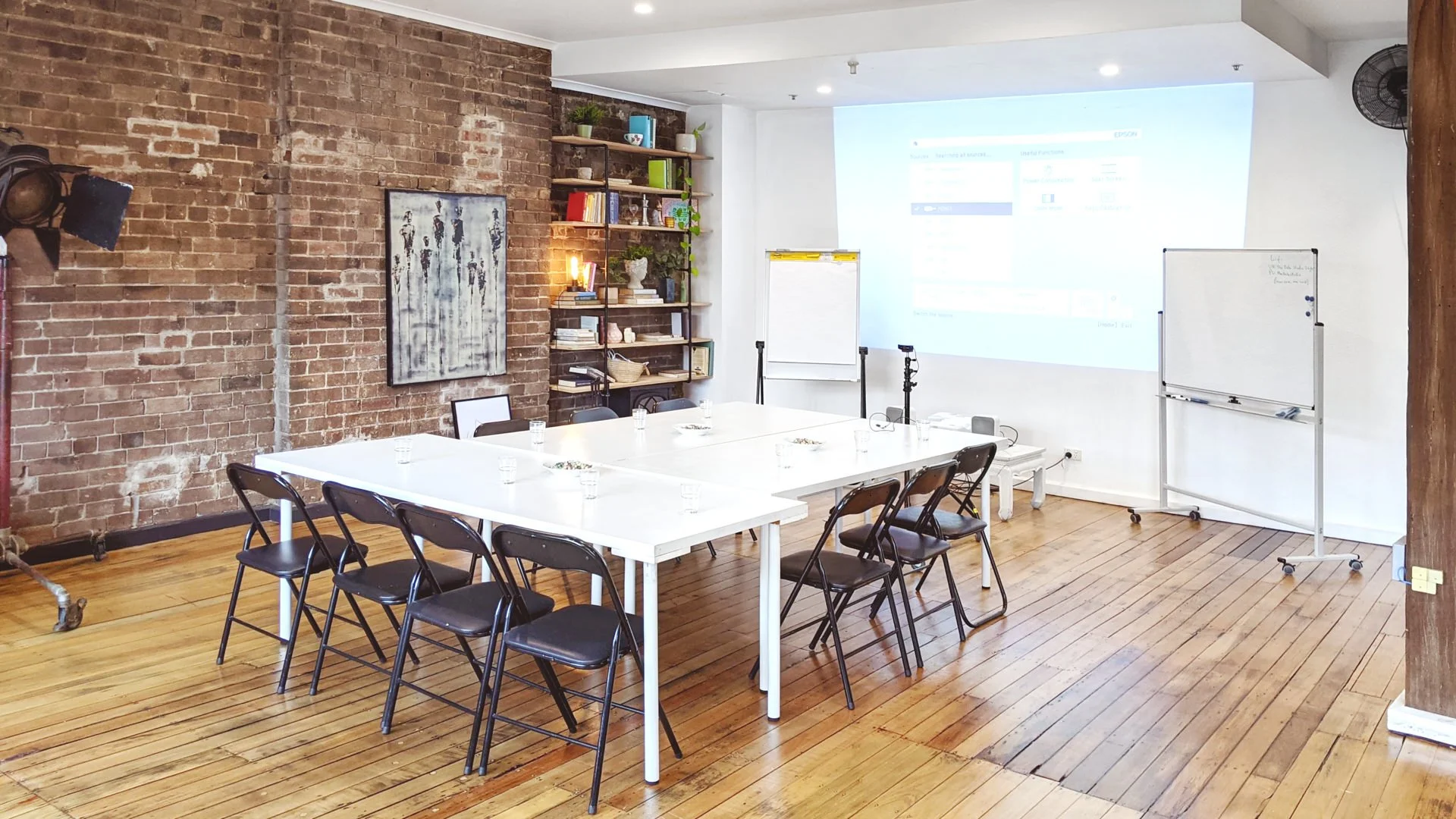 Modern meeting room with tables, folding chairs, whiteboards, projector screen, exposed brick wall, and wood floors