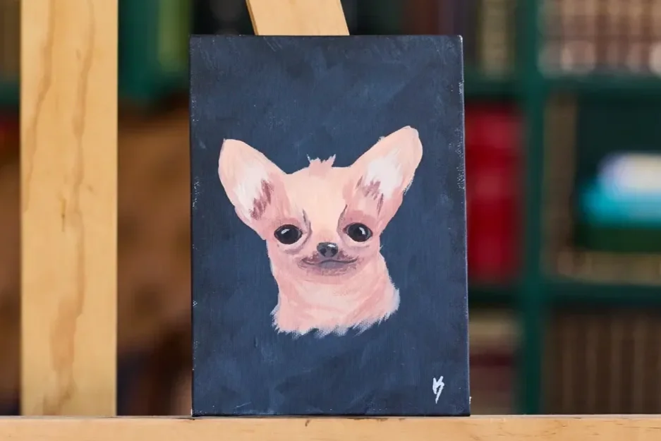 Small painting of a cream Chihuahua with big ears on a charcoal canvas, shown close up on a wooden easel ledge.