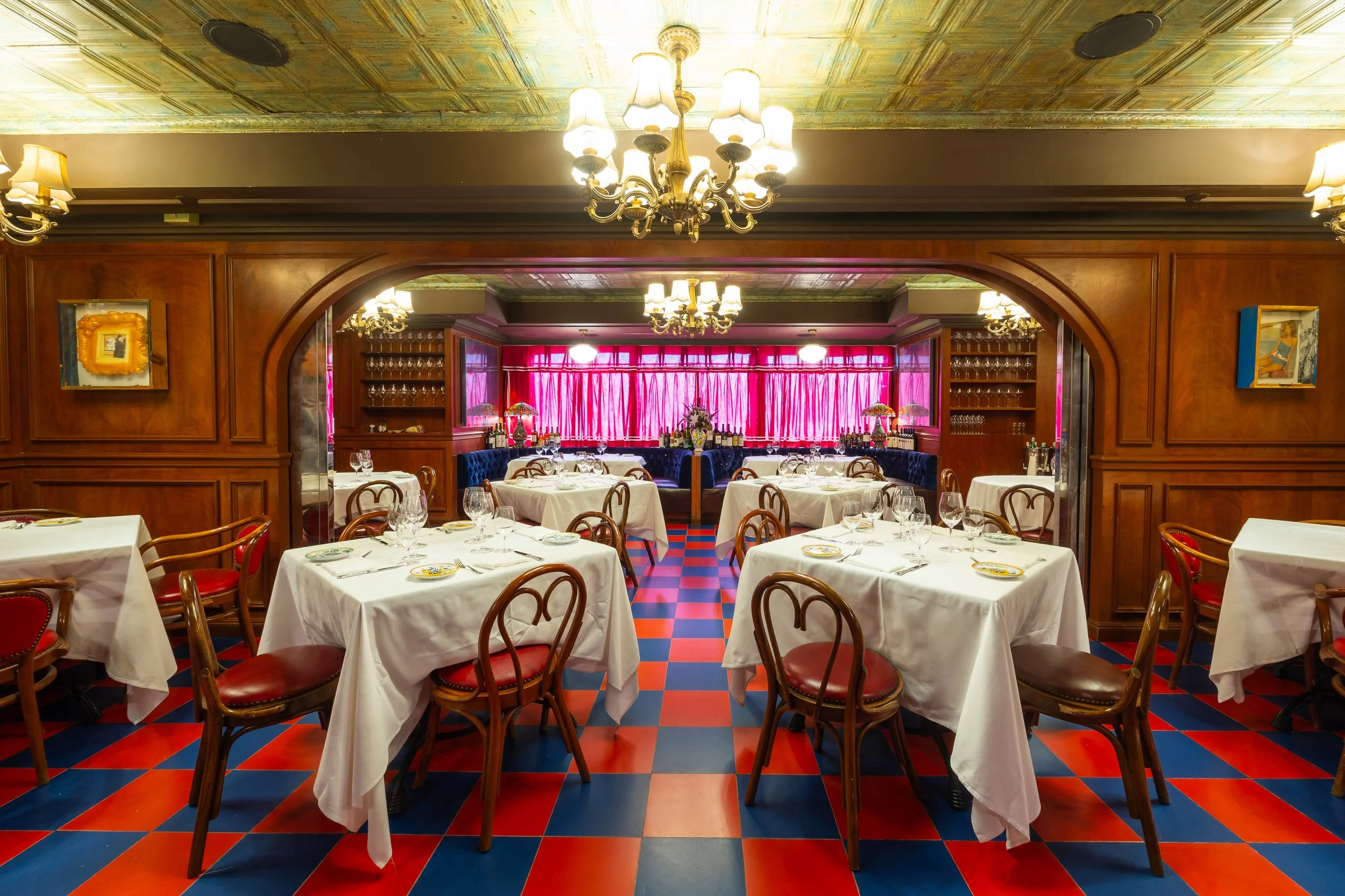 Elegant dining room with white tablecloth tables, classic bentwood chairs, chandeliers, rich wood paneling, and a red-and-blue tiled floor creating a vintage atmosphere.