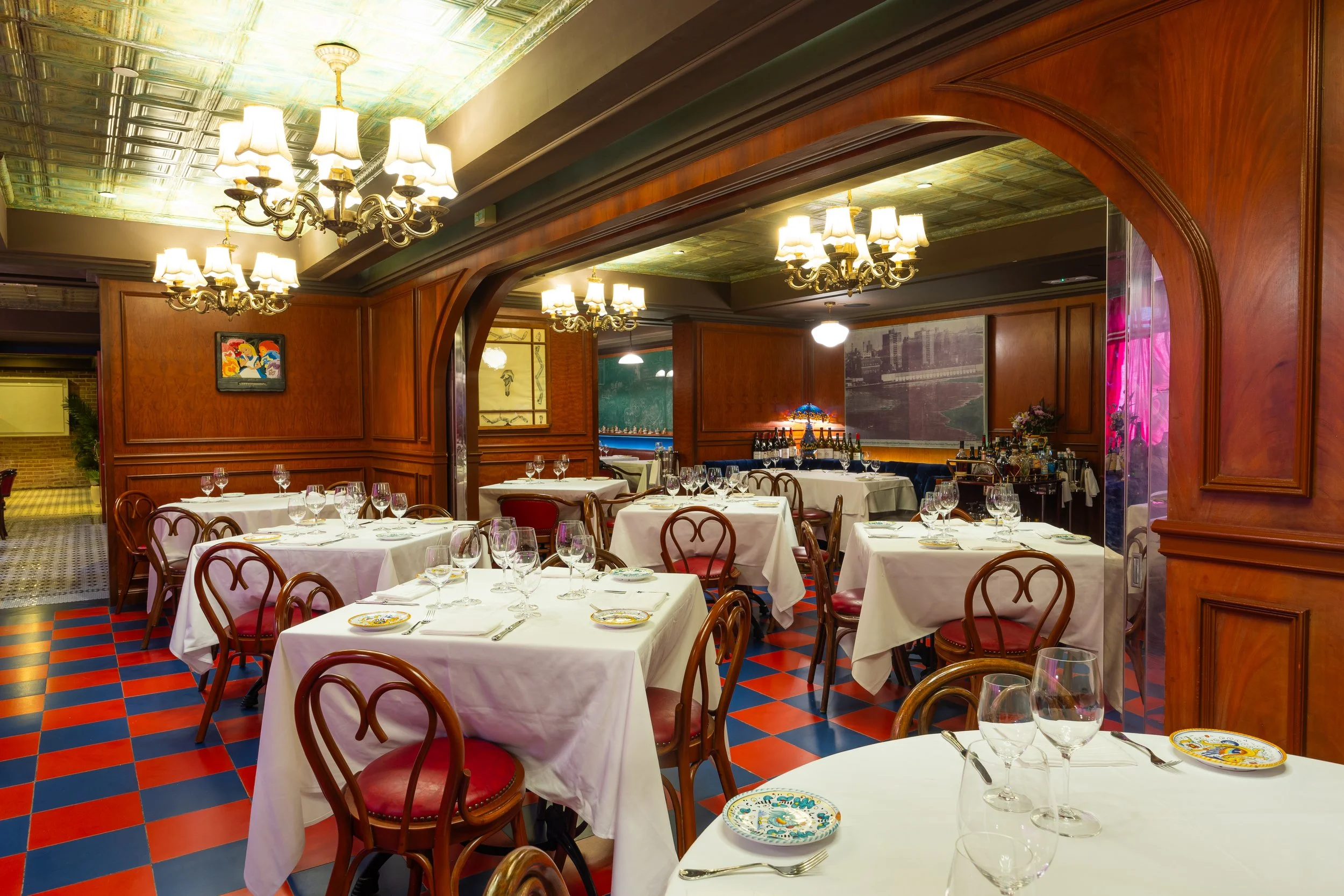 Elegant dining room with white tablecloth tables, classic bentwood chairs, chandeliers, rich wood paneling, and a red-and-blue tiled floor creating a vintage atmosphere.