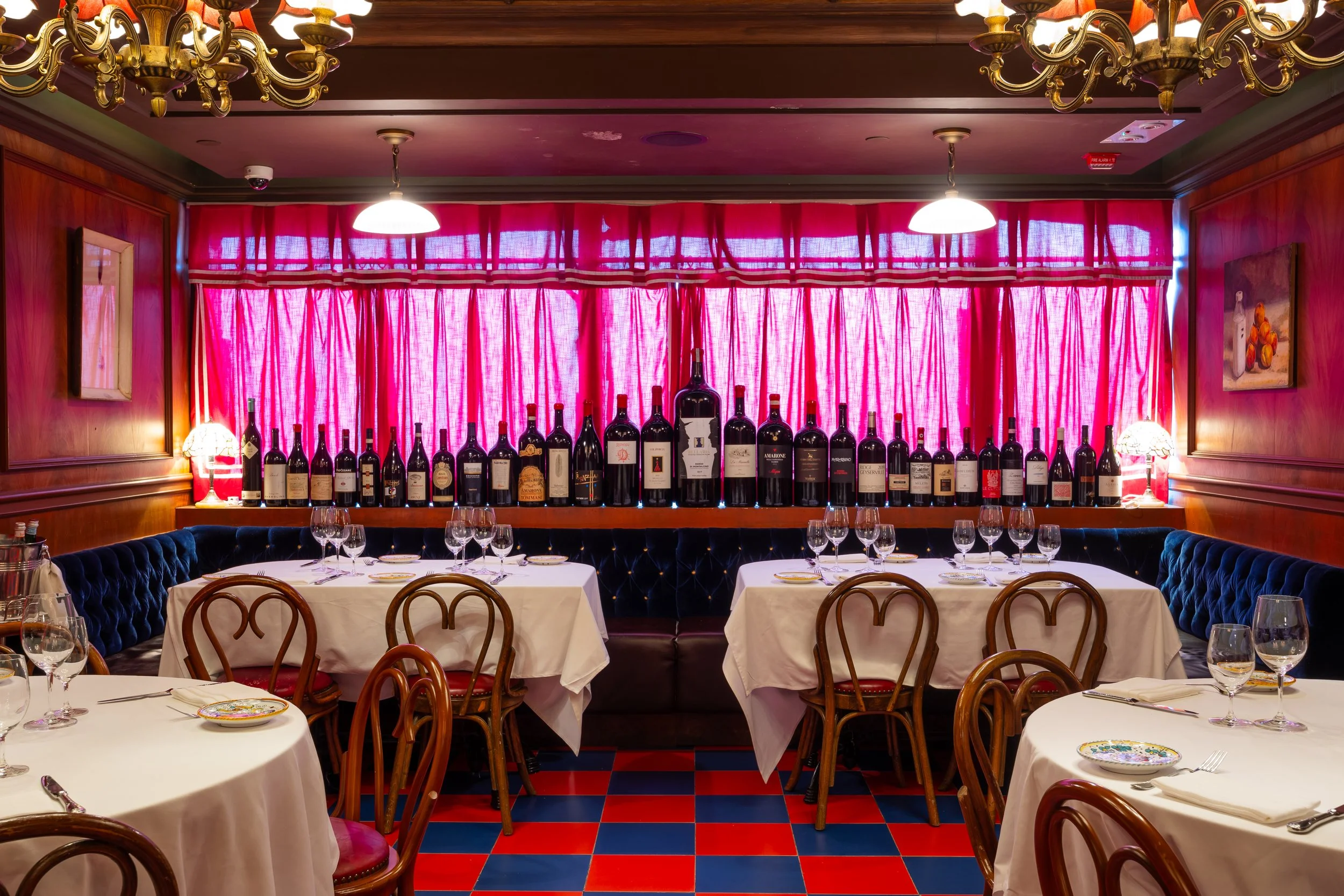 Intimate dining room with white tablecloth tables, bentwood chairs, a blue velvet banquette, red curtains, and a display of wine bottles creating a classic, cozy ambiance.