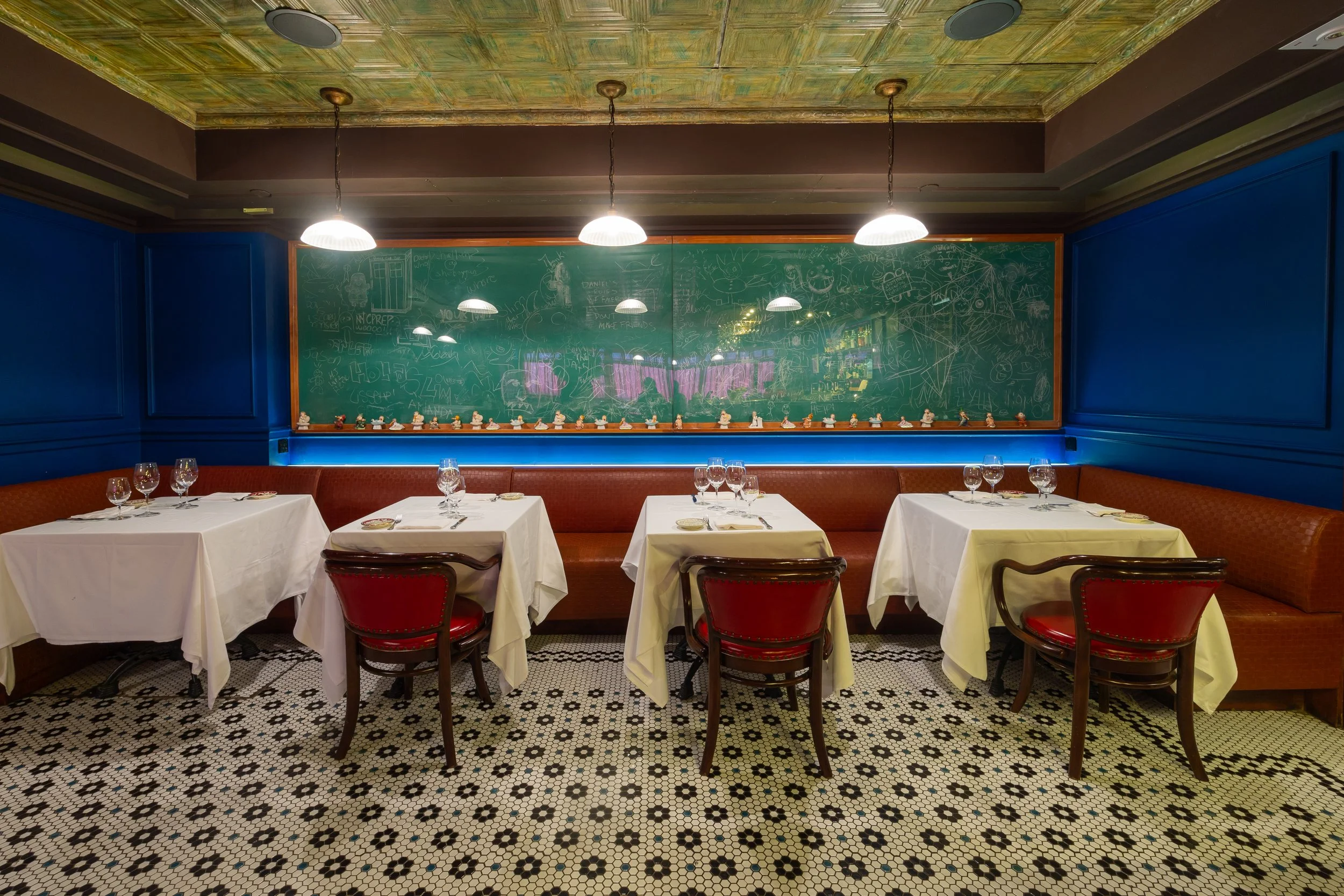 Cozy lounge with white tablecloth tables, red leather banquette and chairs, patterned tile floor, and a chalkboard-style art wall