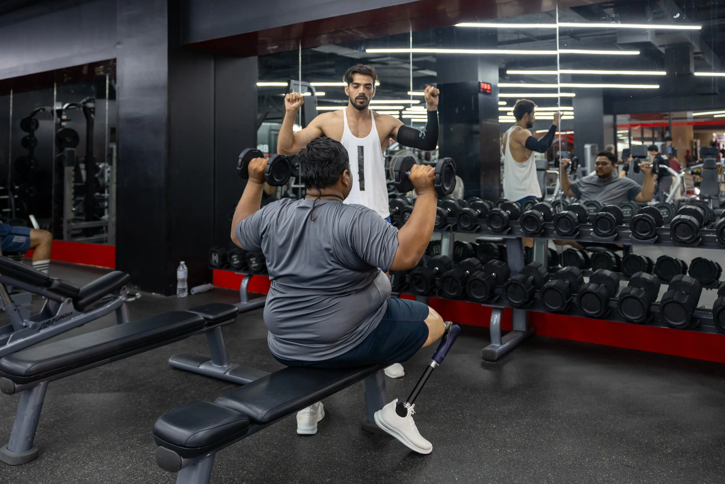 a seated person with a prosthetic leg is doing overhead presses, while a friend/coach demonstrates the. movement standing