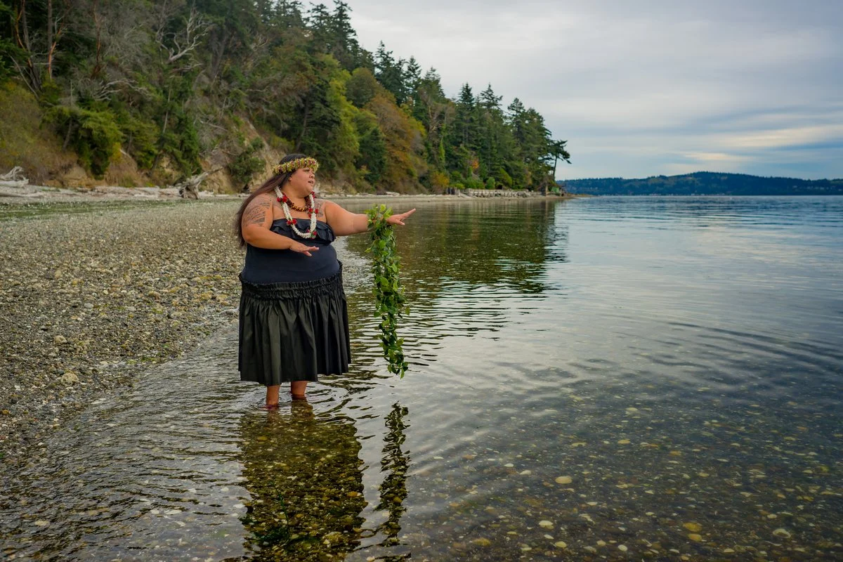 woman in a black dress does a hula dance standing in shallow water on a rocky shore