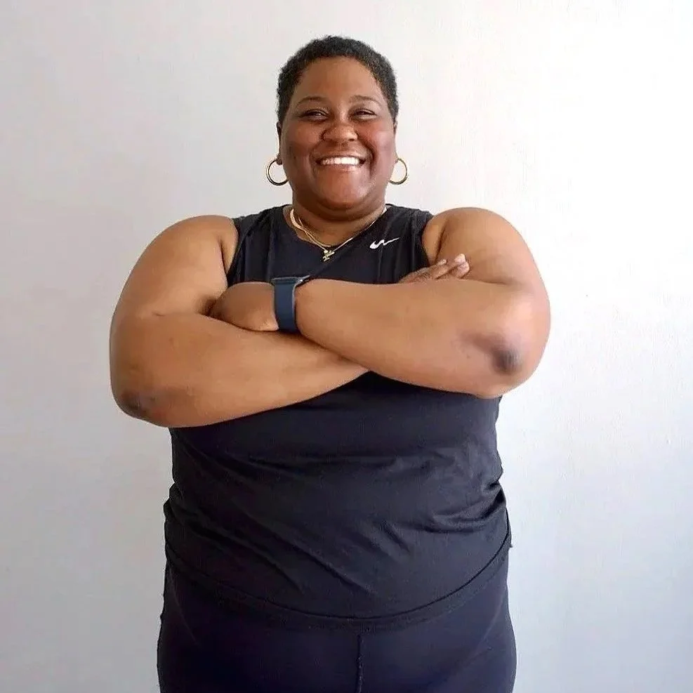 Coach Andrea is a plus size Black woman with short dark hair. She is standing with a smile and her arms crossed