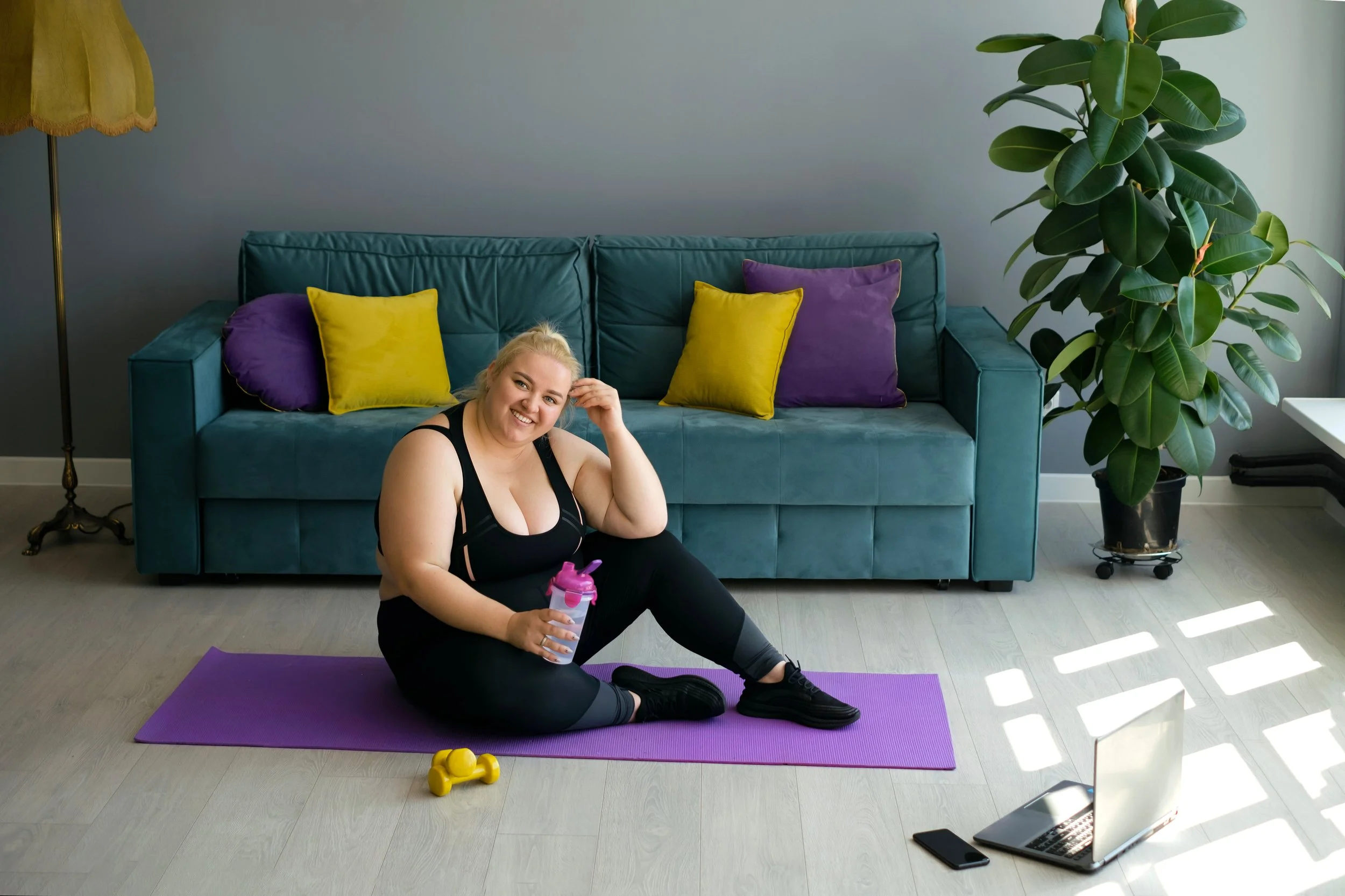 A woman sitting on a yoga mat in a living room, holding a water bottle and smiling, with a laptop, smartphone, yellow dumbbells, and workout clothes nearby.