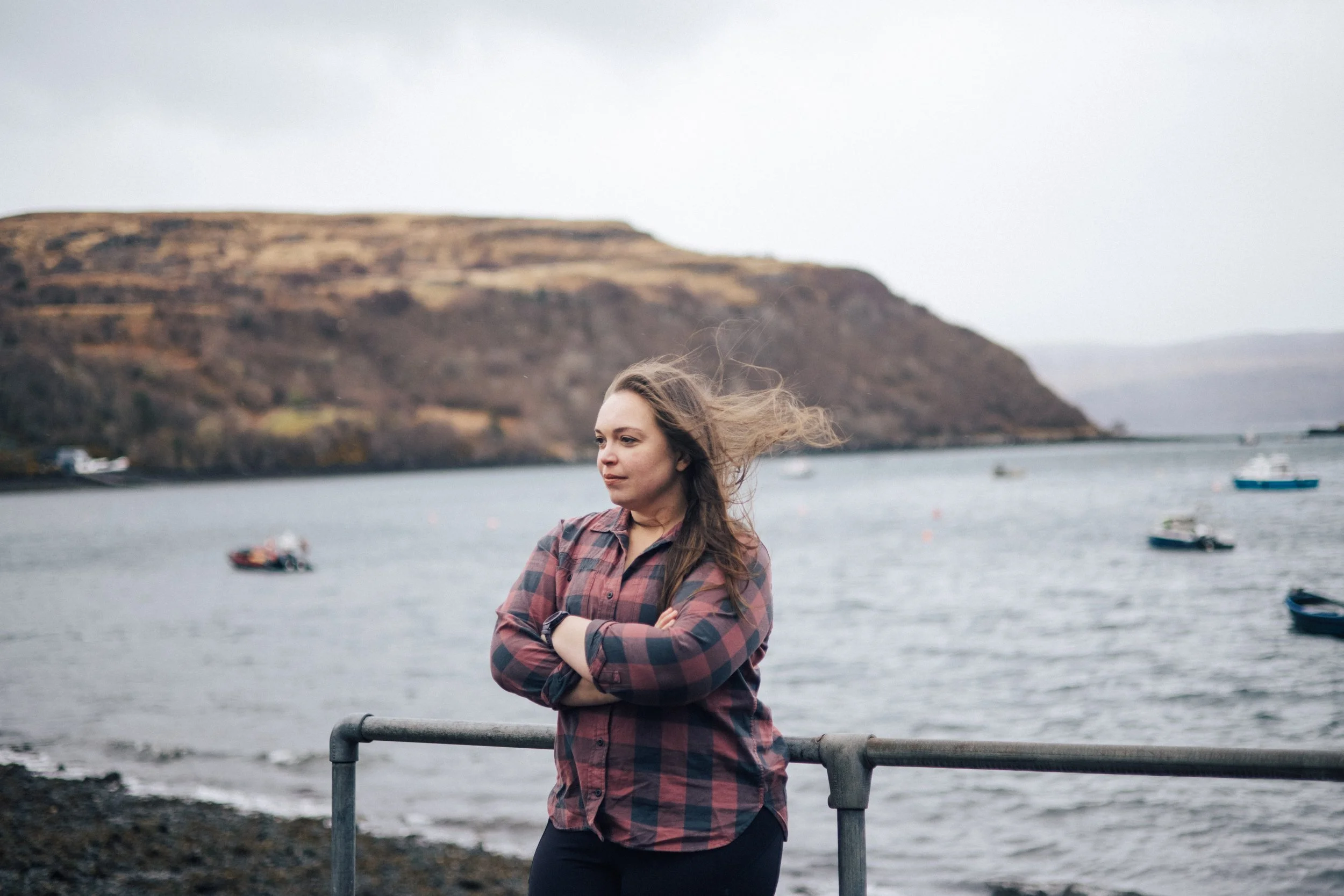 Sarah Petty standing against a railing near Portree Harbor, with boats and hills in the background