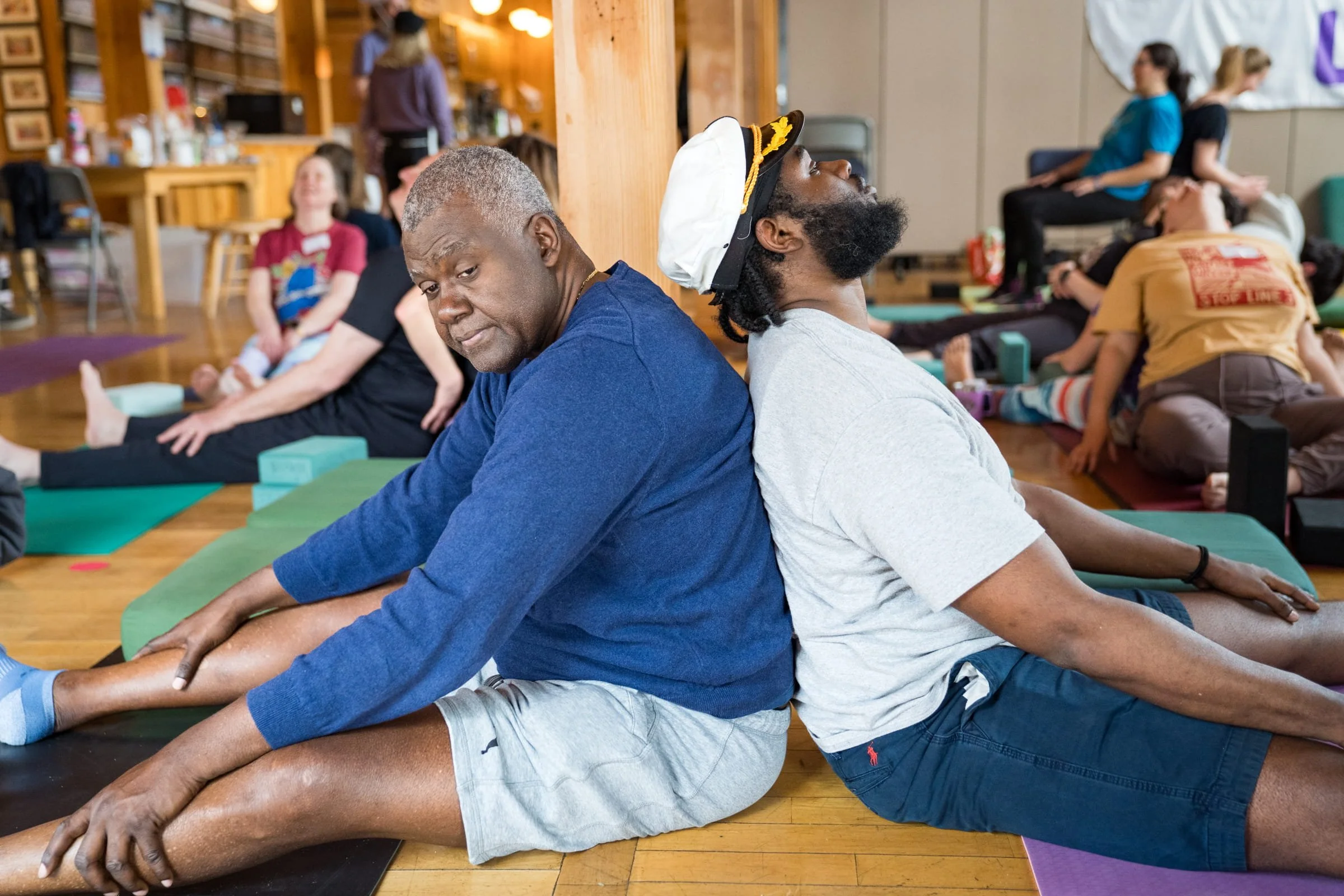 Two BIPOC men sit back to back doing yoga, one is a caregiver for the other