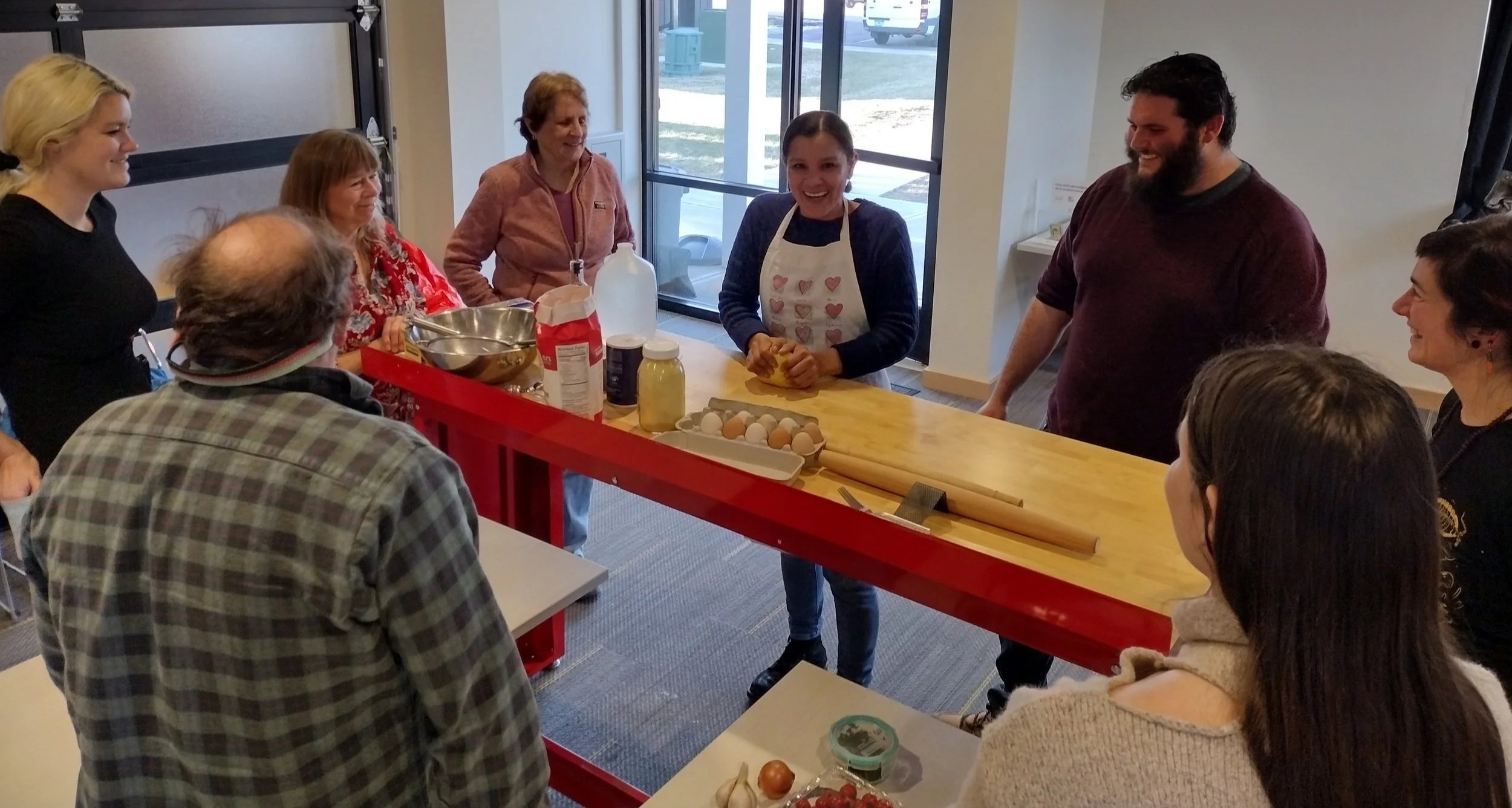 8 adults standing around a long wooden table, with Gloria smiling in the middle as she kneads dough