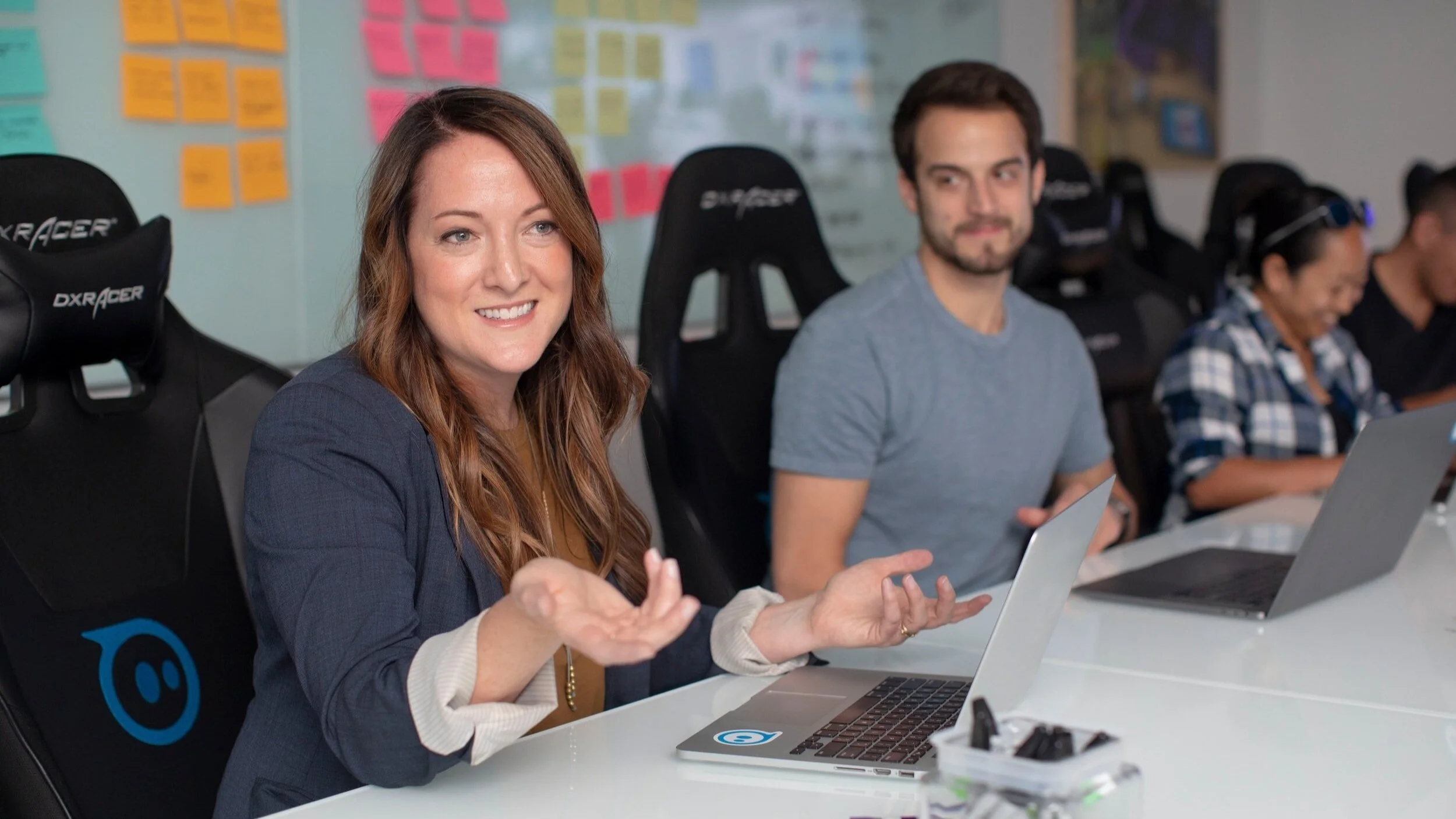 A woman with long brown hair smiling and gesturing with her hands while sitting at a conference table with a laptop in front of her. Two other people, a man with short dark hair and a woman with glasses and a plaid shirt, are also seated at the table, each with a laptop. The setting appears to be a modern office or meeting room with colorful sticky notes on the wall behind them.