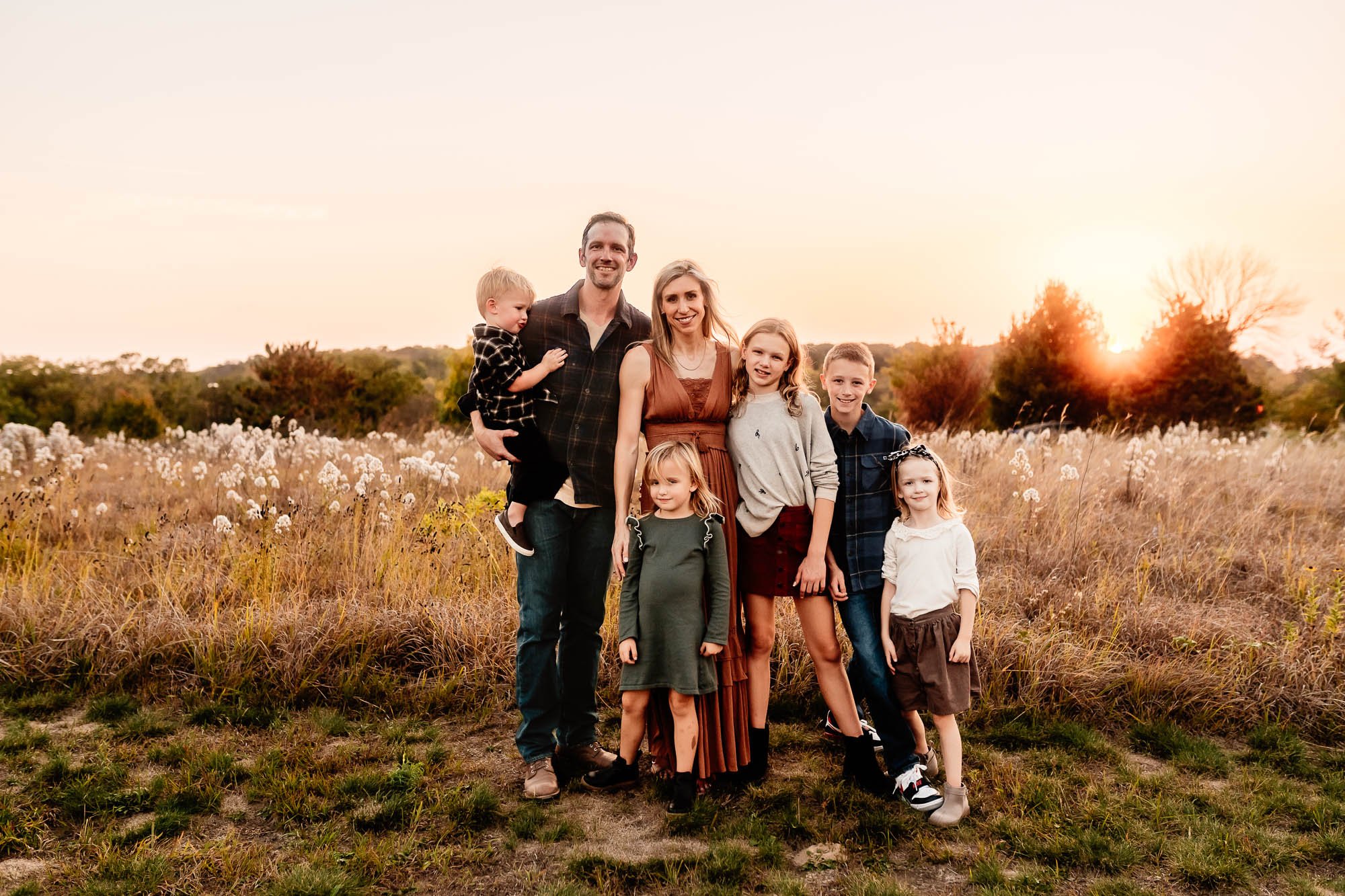 A family of seven posing together in a grassy field at sunset, with warm golden light and autumn tones creating a serene and loving atmosphere.