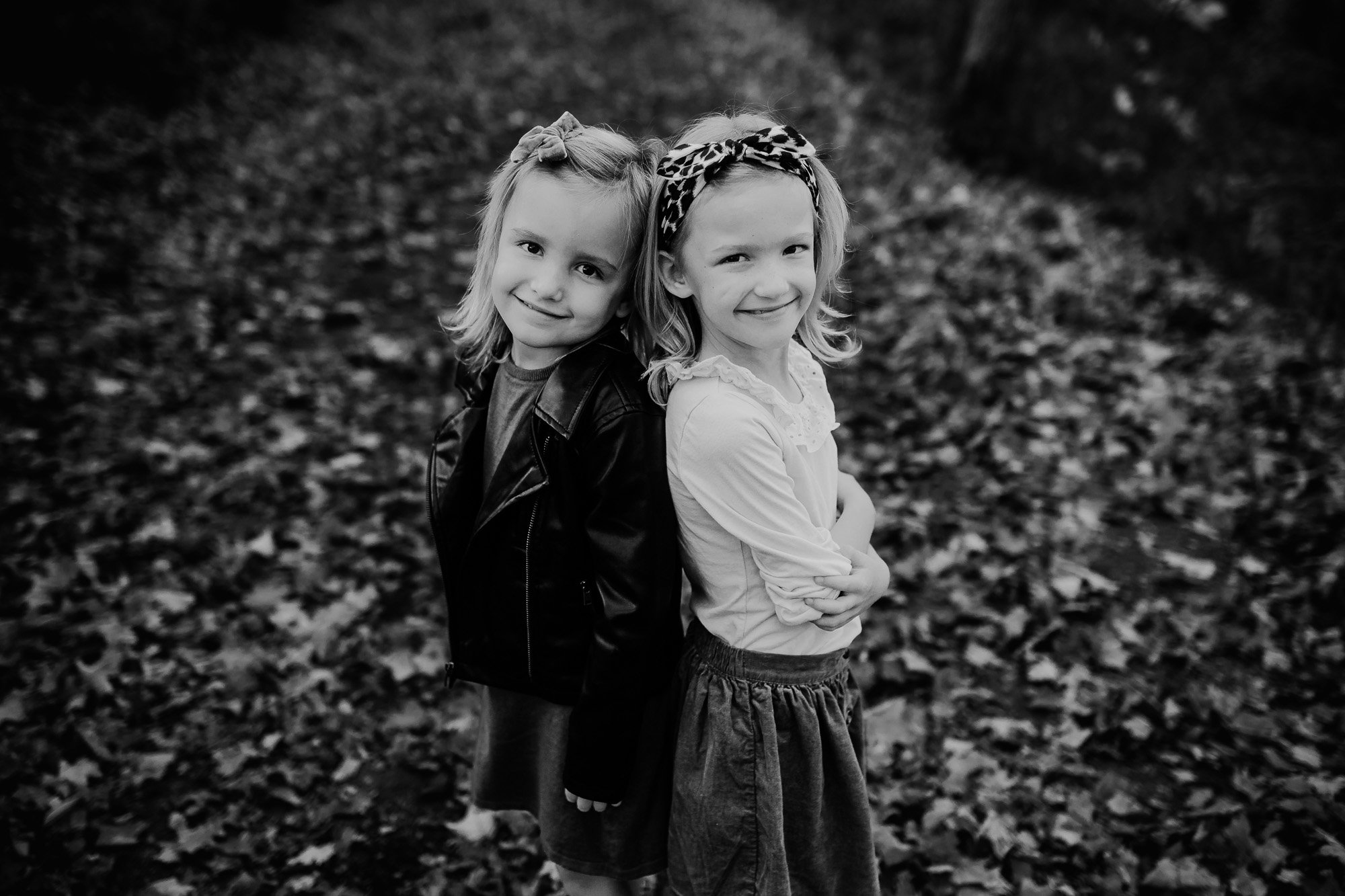 Black and white portrait of two young girls standing back-to-back, smiling sweetly, surrounded by fallen autumn leaves